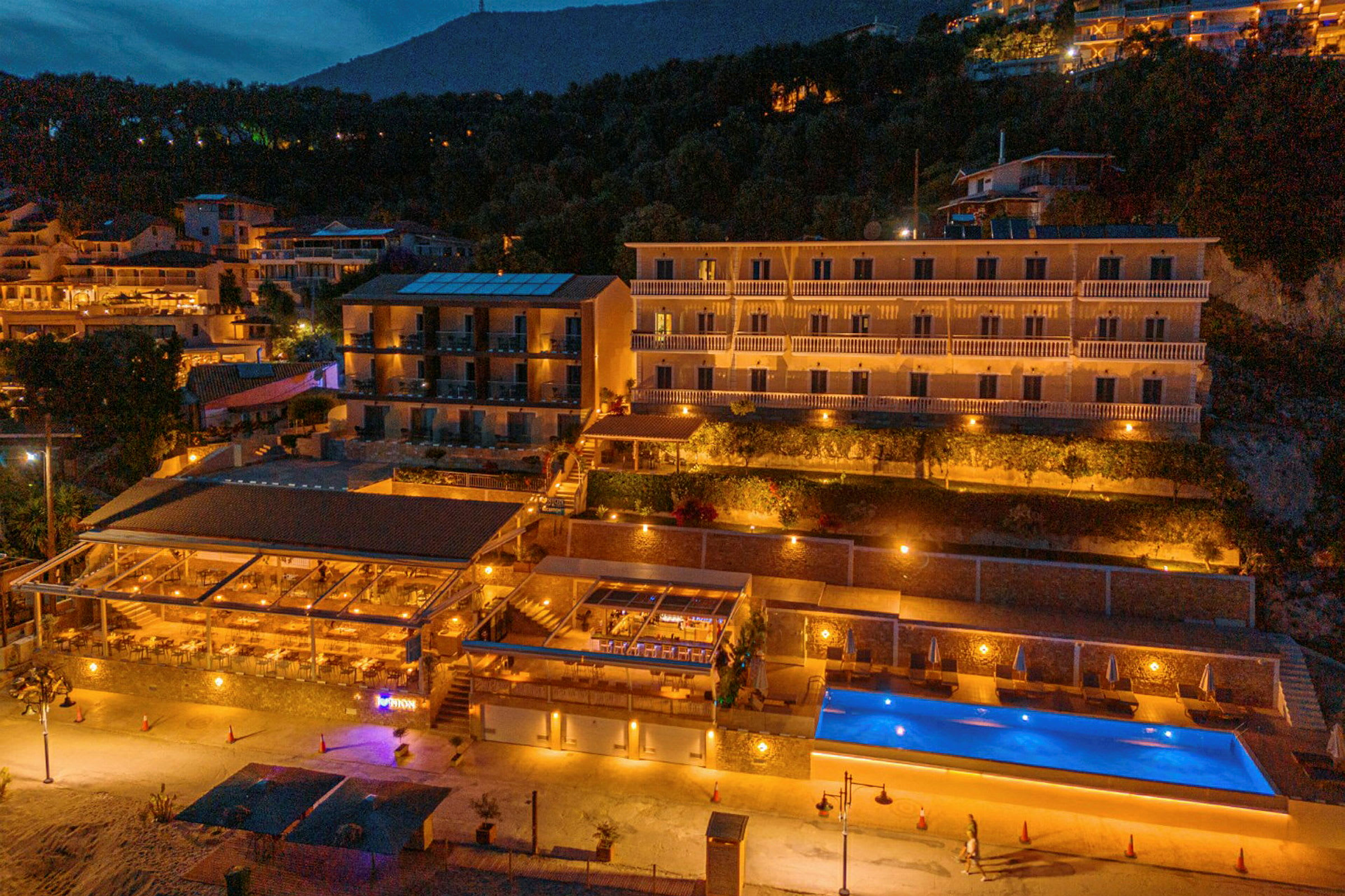 Hotelbygning med pool og restaurant oplyst om aftenen. (AI-genereret tekst)