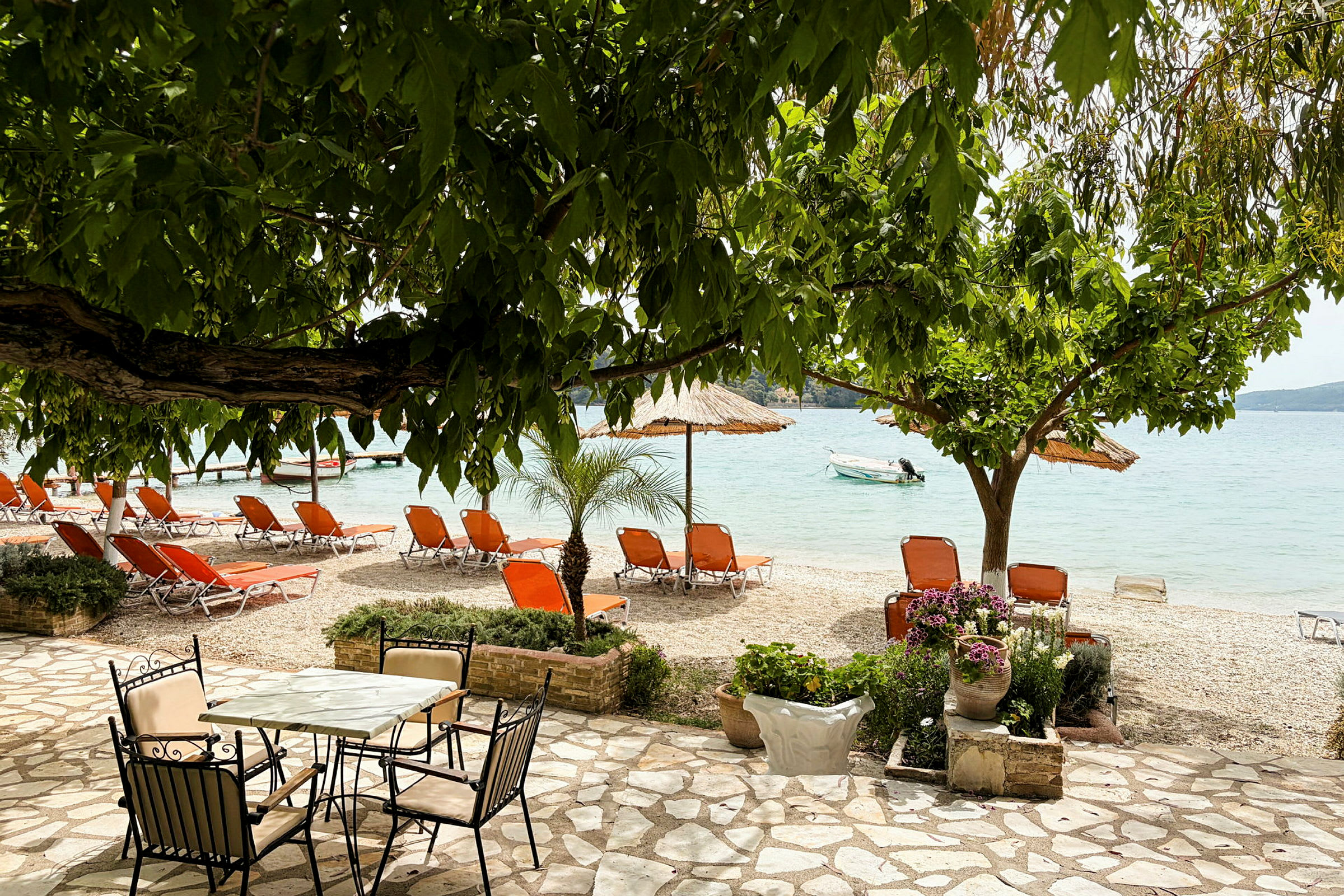 En strand med orange liggestole, parasoller og udsigt over havet. Et stenlagt område med bord og stole samt plantede blomster i forgrunden. (AI-genereret tekst)