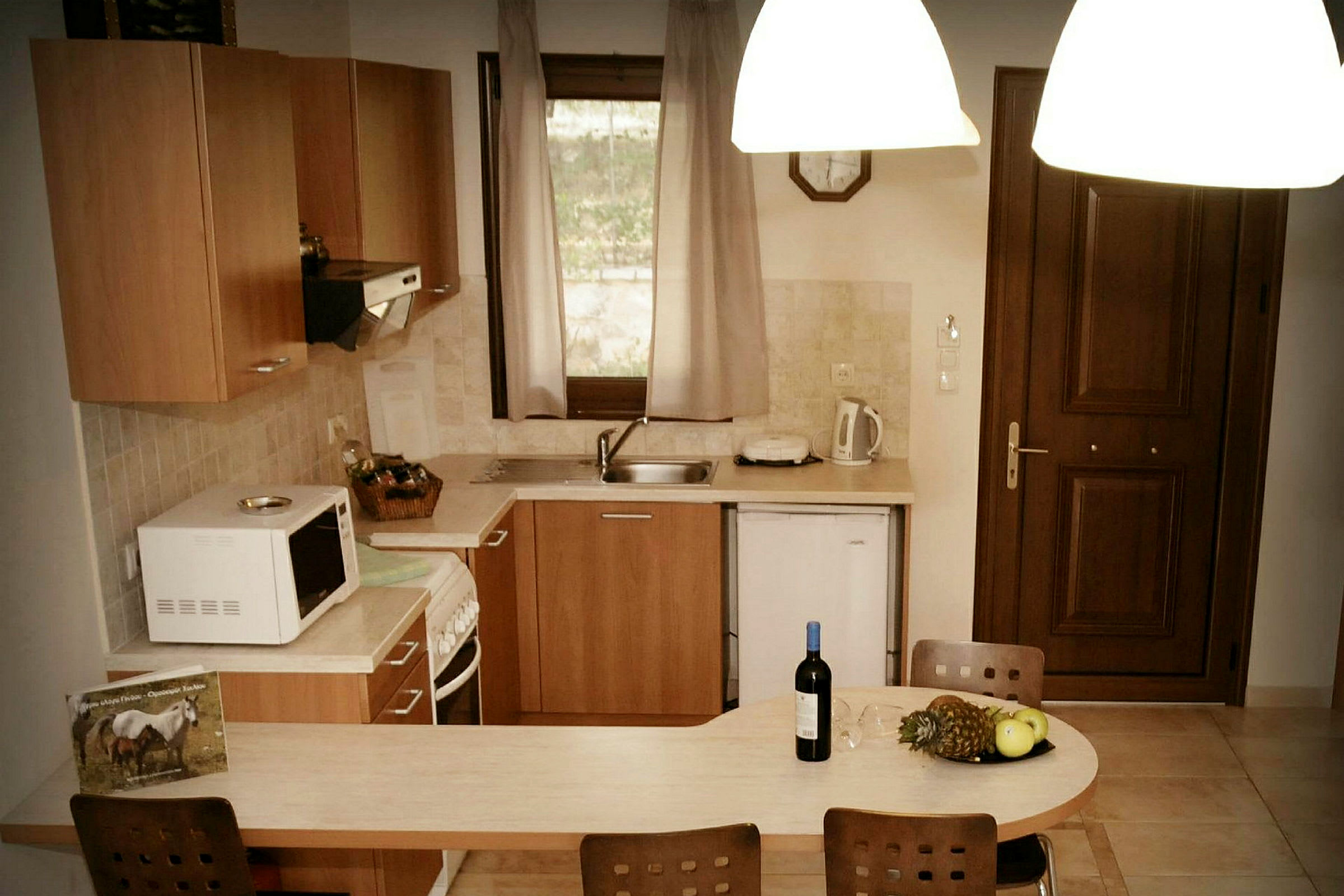 En køkkenområde med håndvask, mikrobølgeovn og køkkenskabe. På køkkenbordet er der en flaske vin og frugt. (AI-genereret tekst)