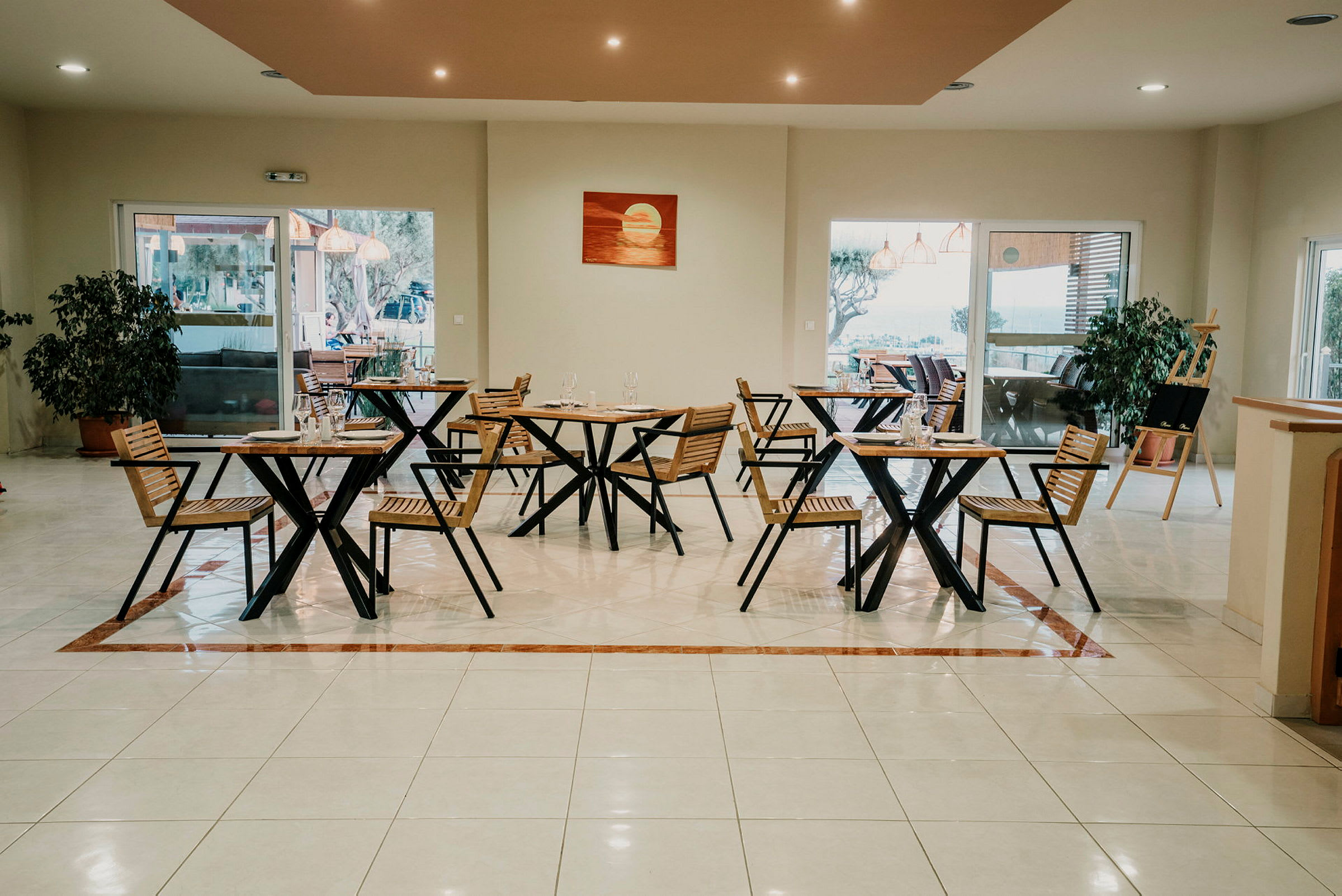 En restaurant med træstole og borde, store vinduer og en indendørs plante på gulvet. (AI-genereret tekst)