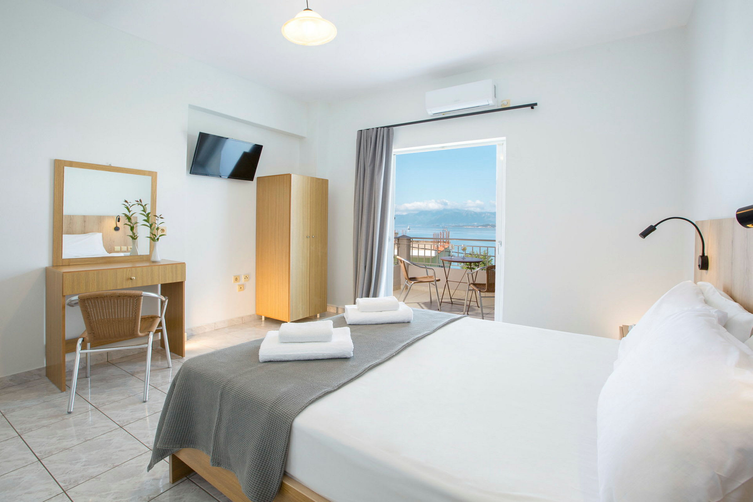 Et lyst hotelværelse med en dobbeltseng, skrivebord, spejl og en altan med udsigt over havet. (AI-genereret tekst)