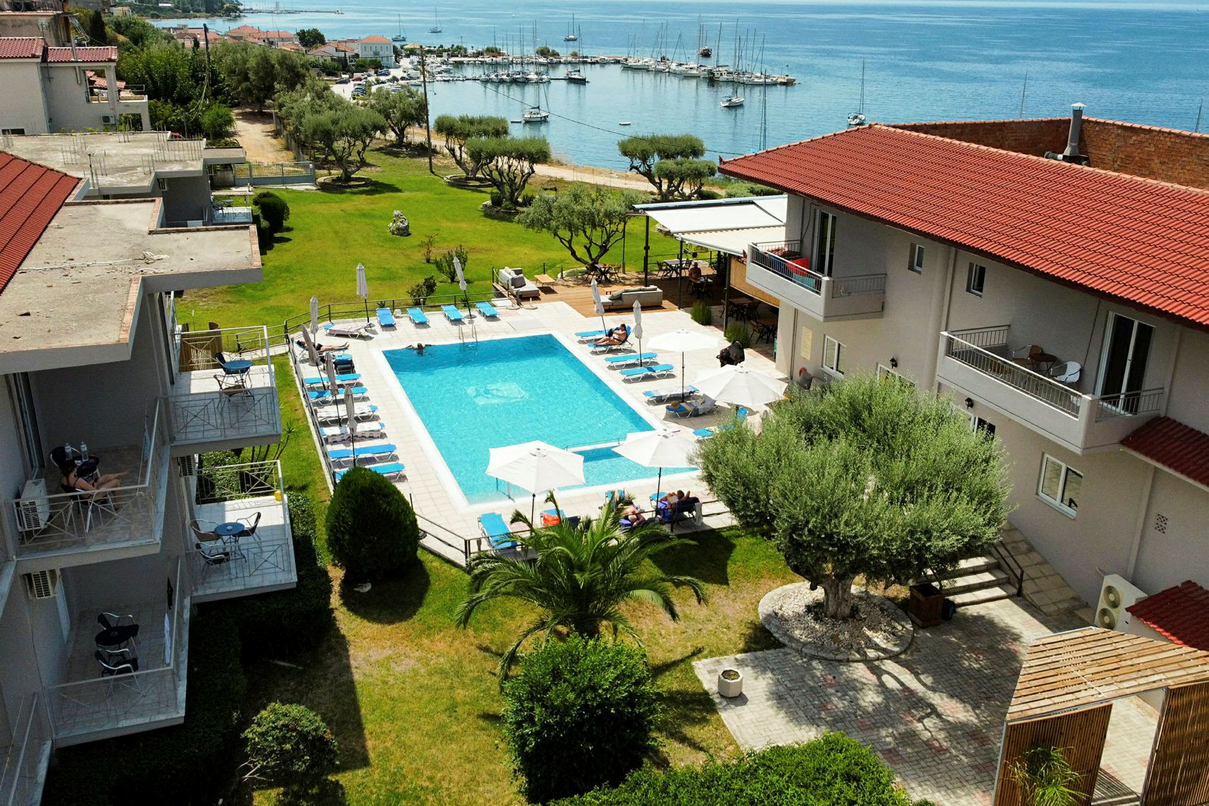 Billede af et hotelområde med en udendørs pool, liggestole og parasoller, samt omkringliggende grønne områder og udsigt mod en havn ved havet. (AI-genereret tekst)