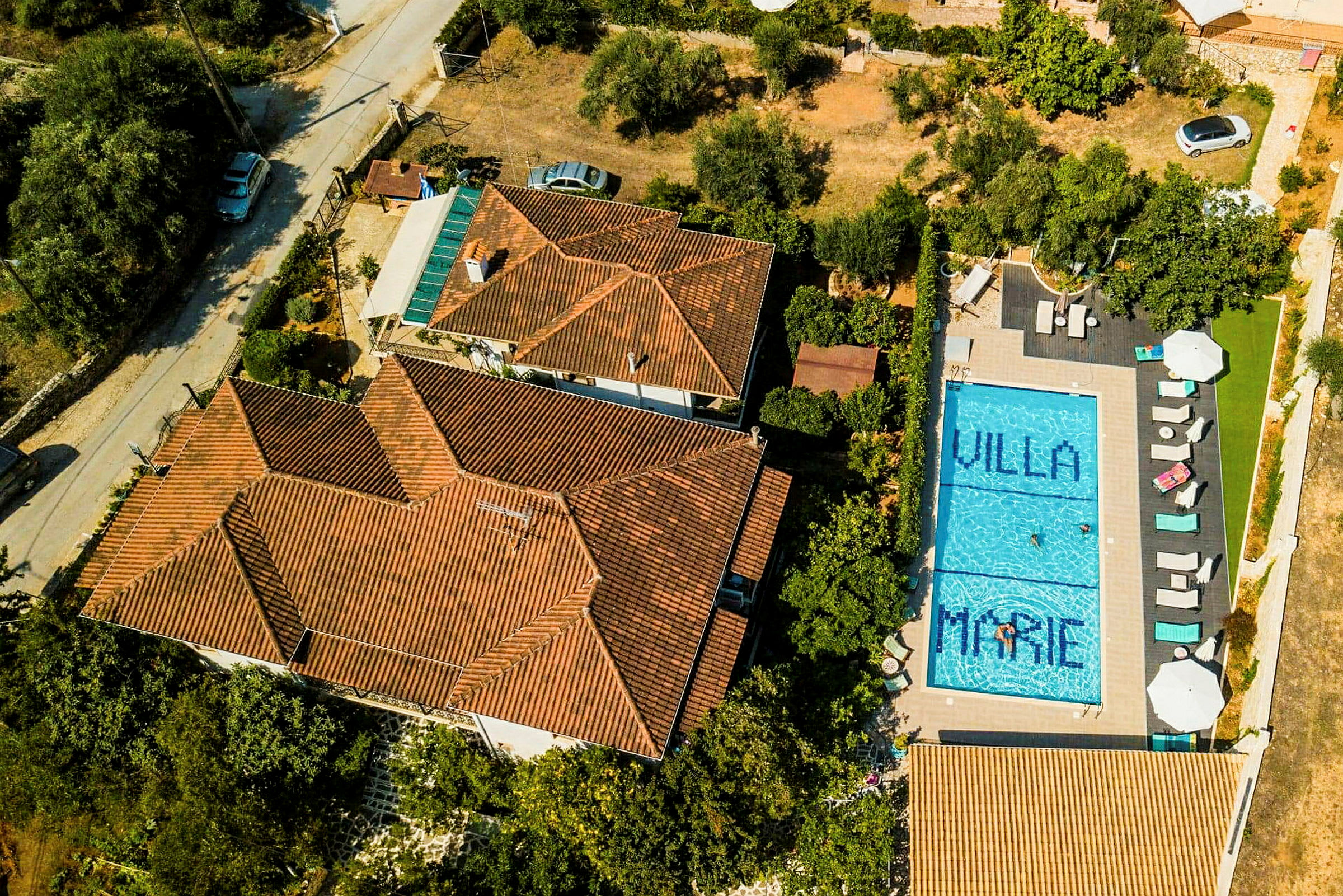 Luftfoto af en villa med orange tegltag og en blå swimmingpool på grunden. Poolen har teksten 'Villa Marie'. (AI-genereret tekst)