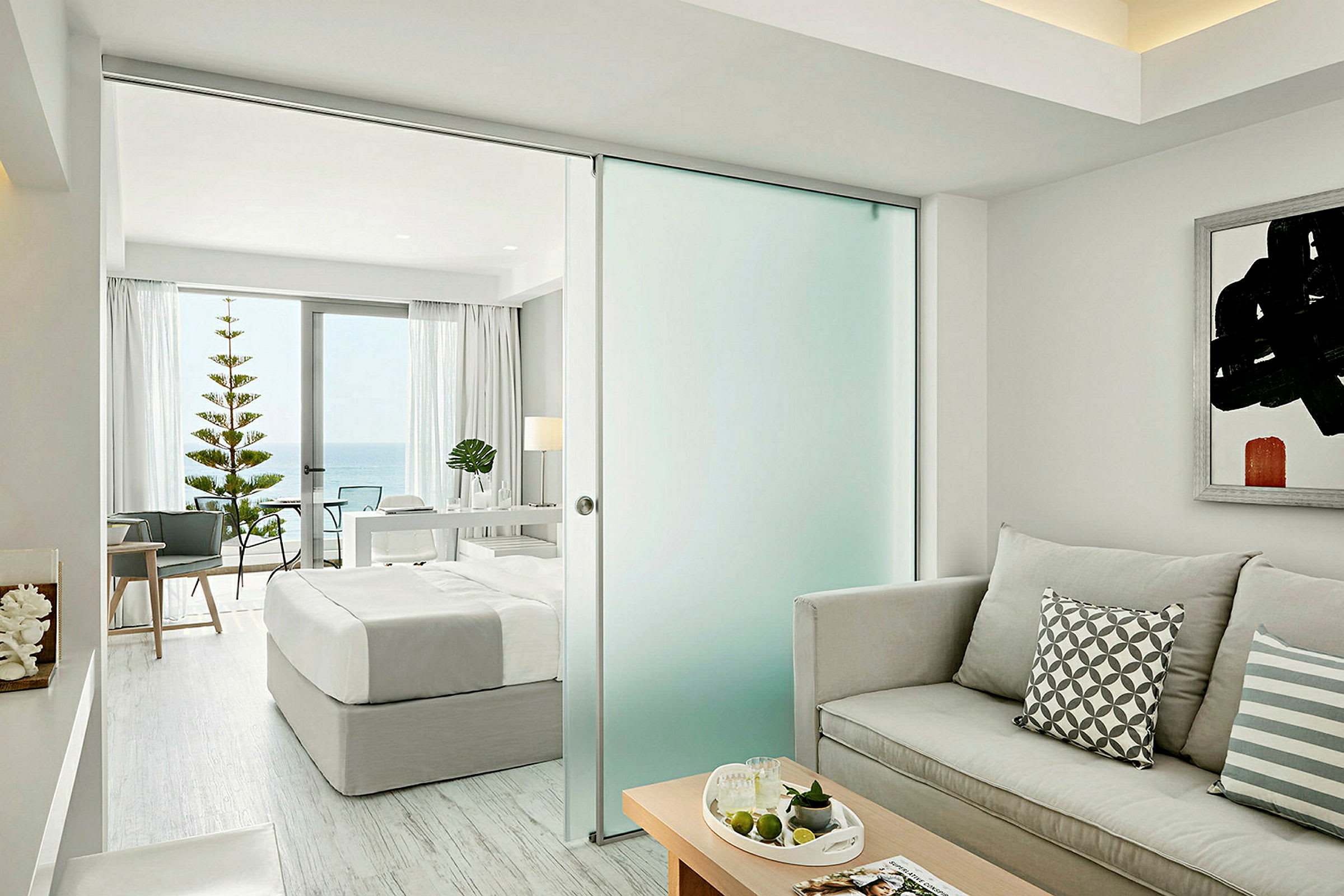 Et lyst og moderne hotelværelse med en seng, sofa og udsigt mod havet gennem store vinduer. (AI-genereret tekst)