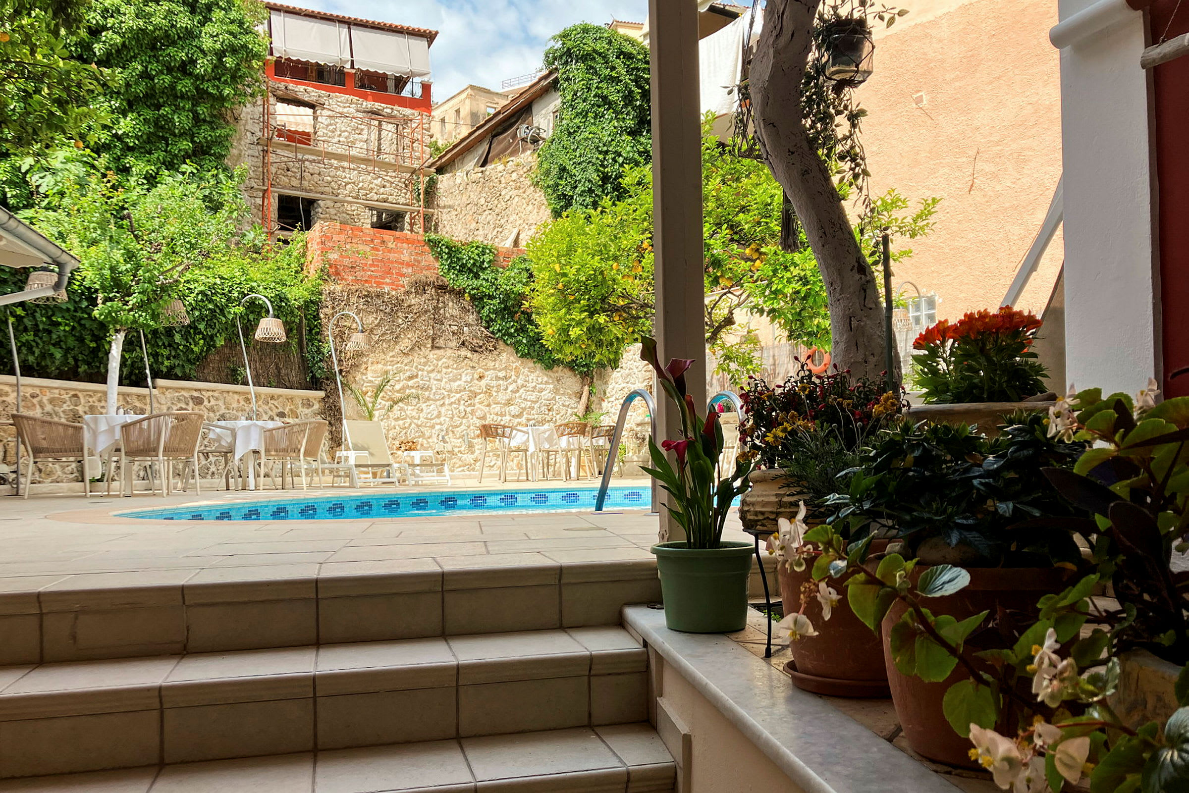 En terrasse med en pool omgivet af borde og stole, grønne planter i potter og en stenmur med vegetation. (AI-genereret tekst)