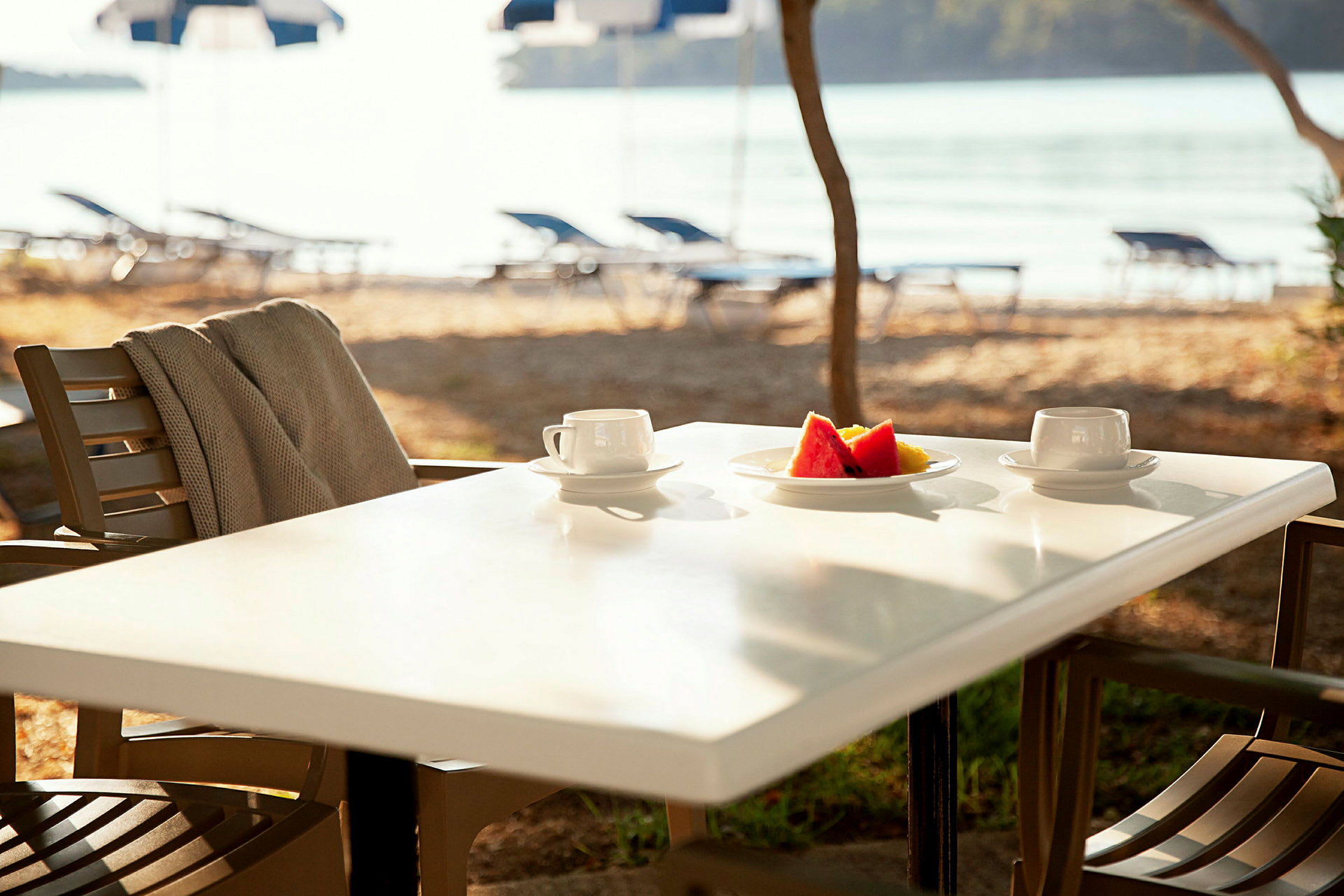 En udendørs servering med et bord med to kaffekopper og en tallerken med frugt. Baggrunden viser strand og vand. (AI-genereret tekst)