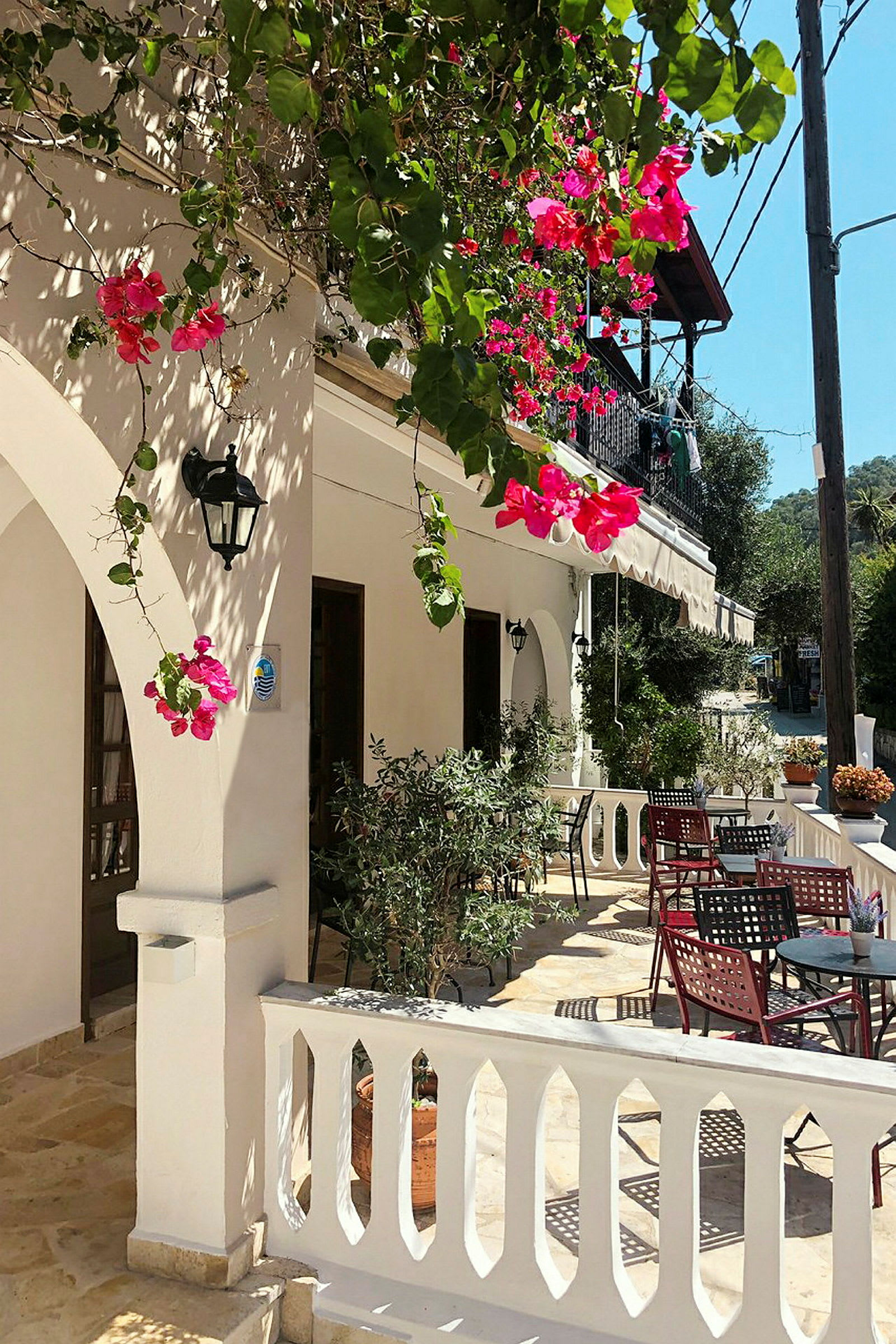 En solrig terrasse med hvide balustrader og borde omgivet af blomstrende bougainvillea. (AI-genereret tekst)
