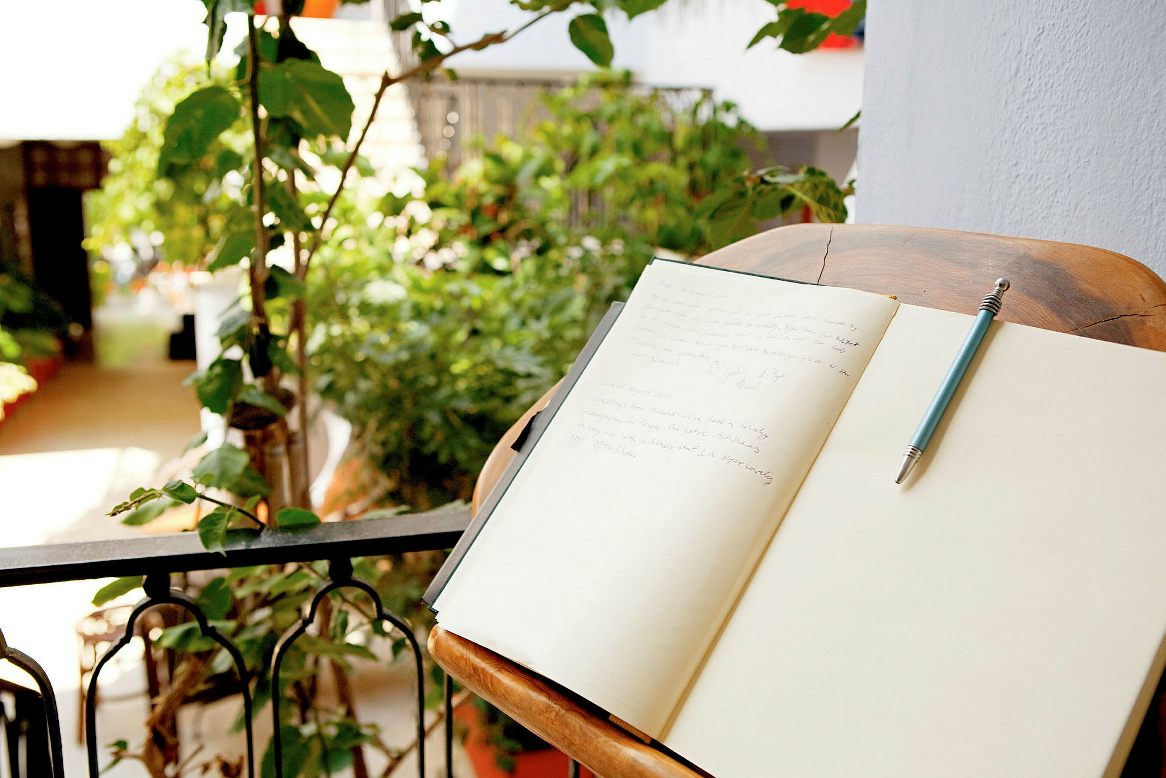 Et skrivebord med en åben notesbog og en pen ved siden af, placeret på en balkon med planter i baggrunden. (AI-genereret tekst)