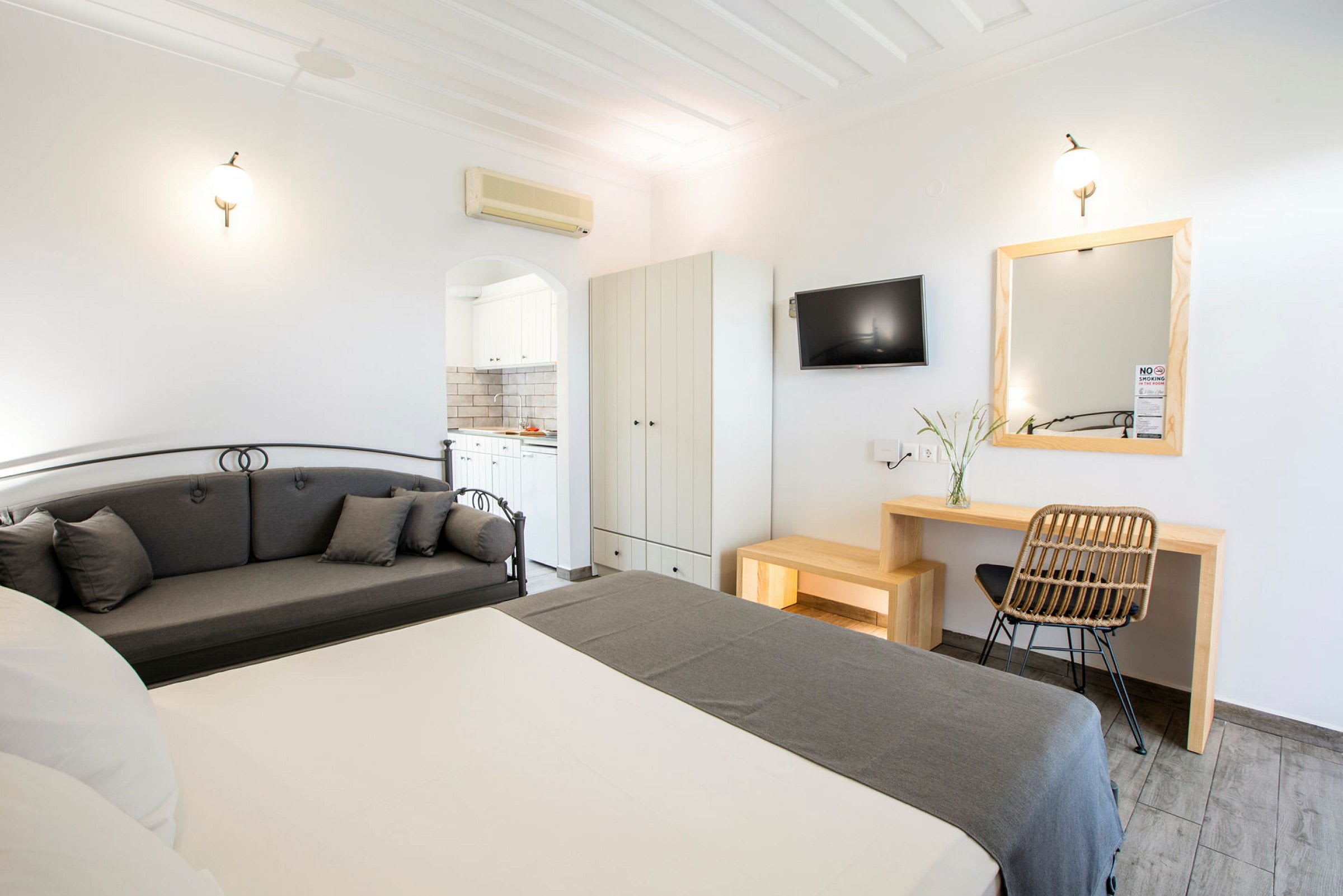 Et hotelværelse med seng, grå sofa, skrivebord, vægmonteret TV og garderobe. Et badeværelse kan ses gennem en døråbning. (AI-genereret tekst)