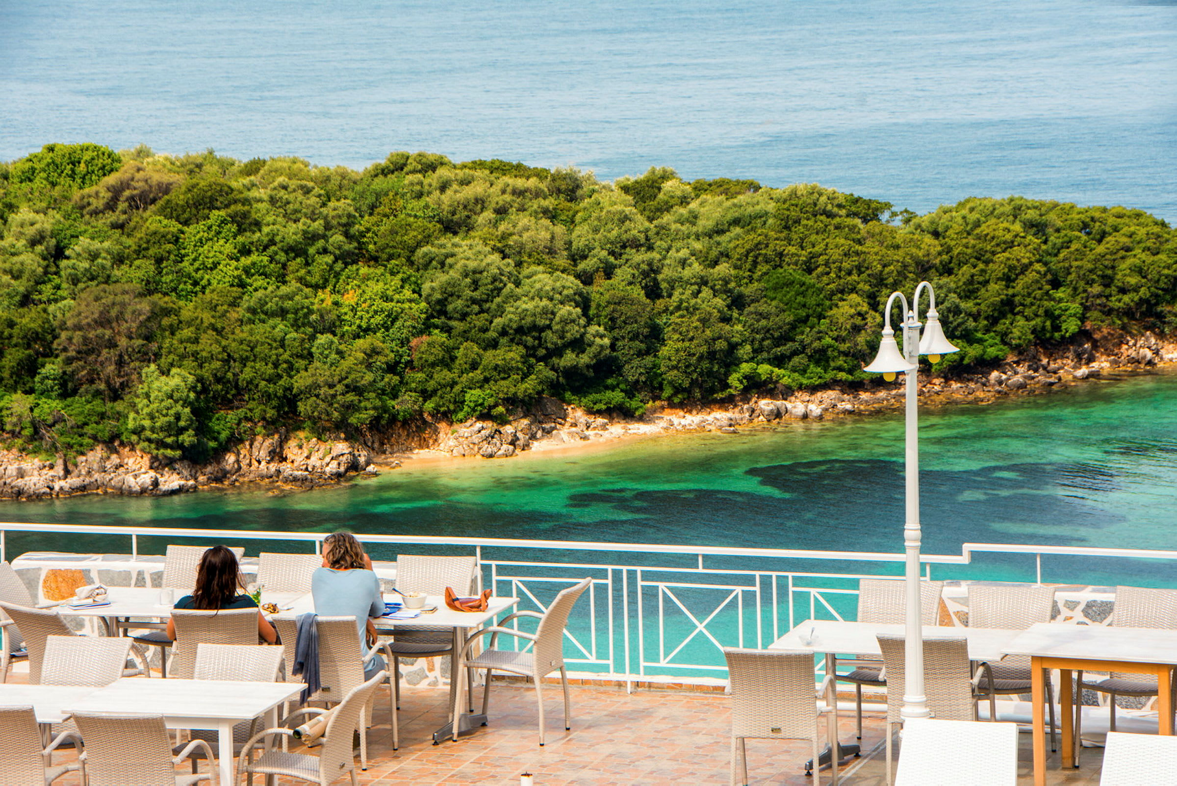 To personer sidder ved borde på en terrasse med udsigt over havet og en frodig kystlinje. (AI-genereret tekst)