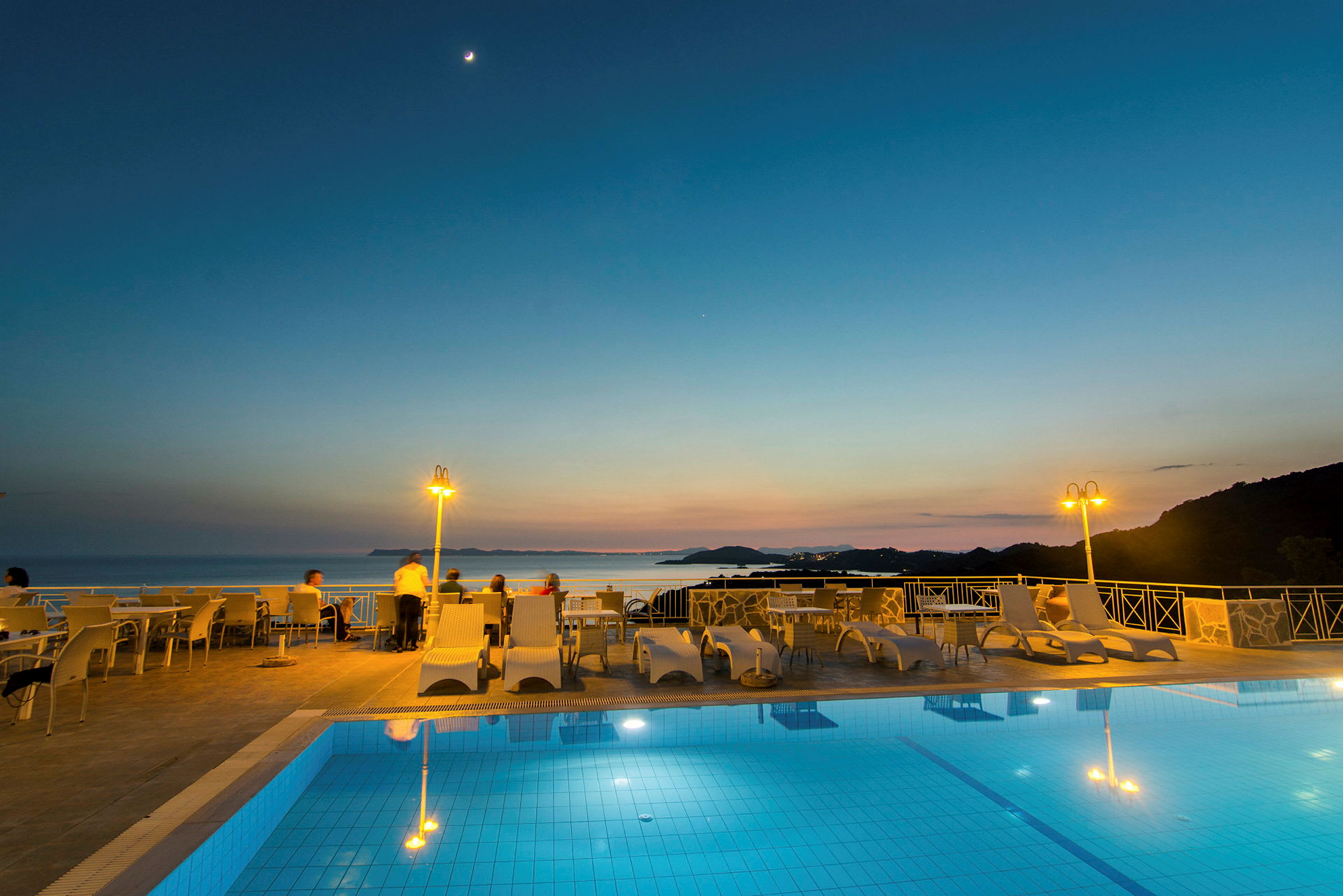 Udsigt over en pool og siddepladser med havet og solnedgangen i baggrunden. (AI-genereret tekst)