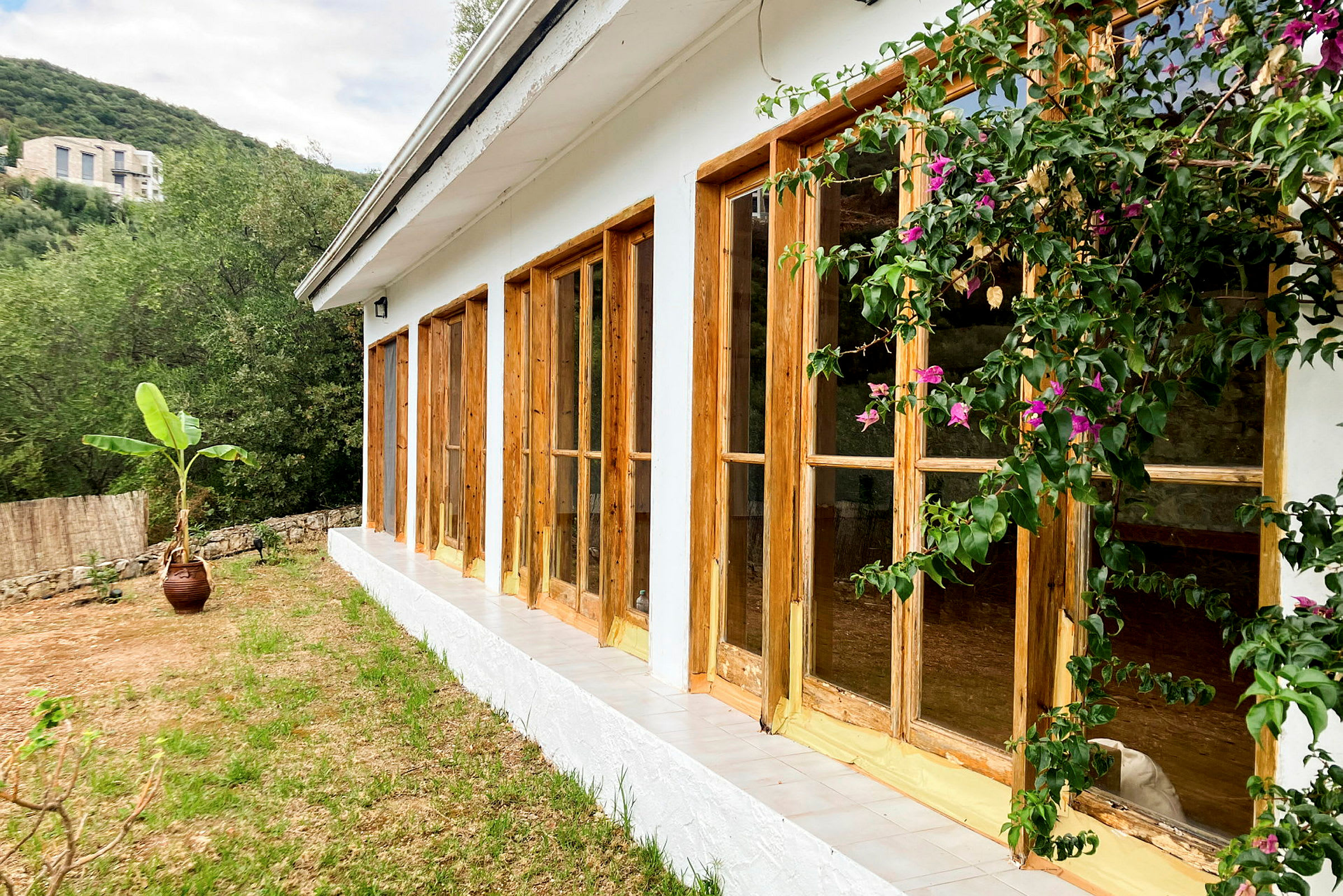 Billede af et hus med hvide vægge og vinduer med trærammer, omgivet af grønt og blomster. (AI-genereret tekst)