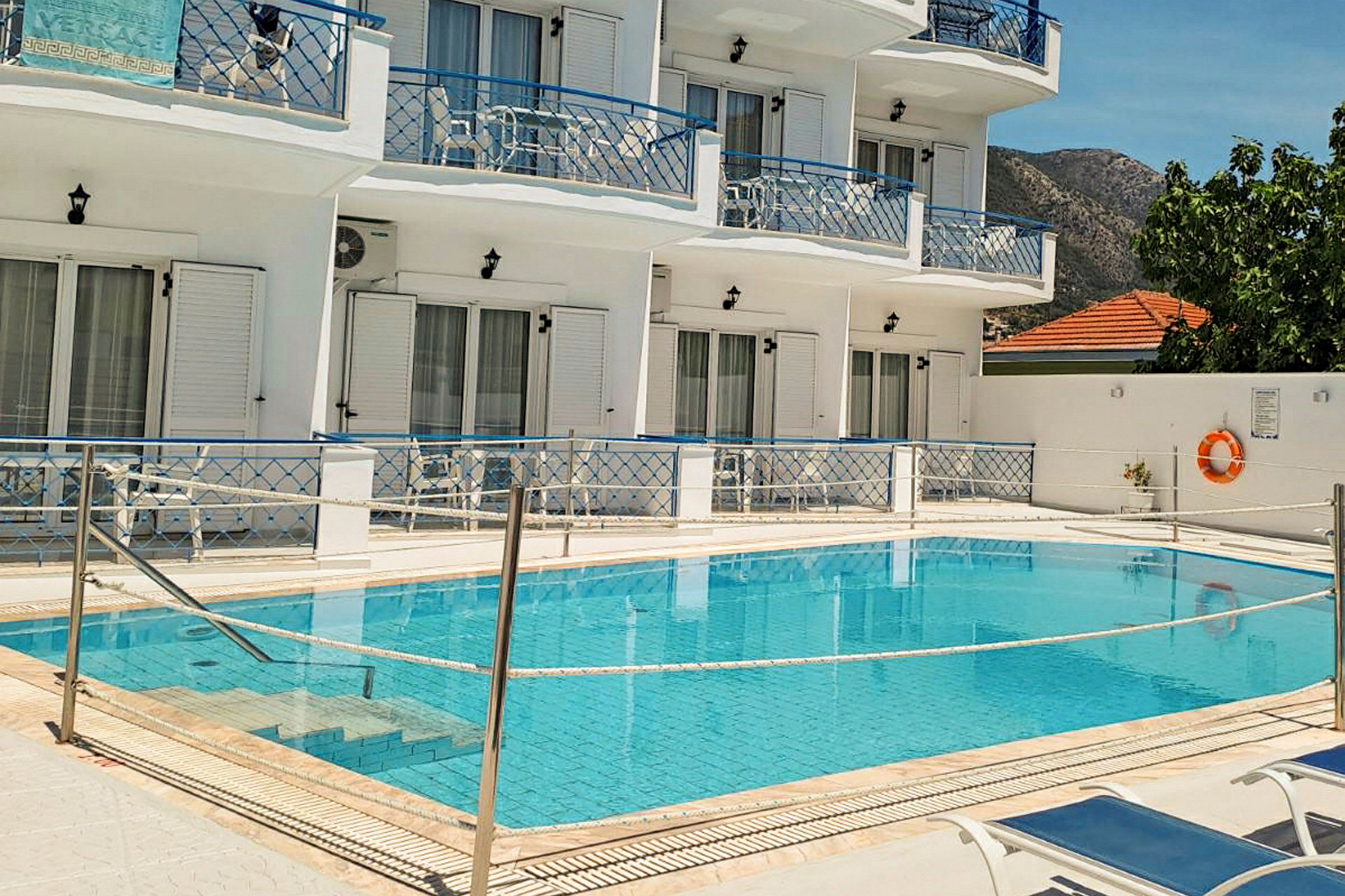 Billede af en pool foran et hvidmalet hotel med blå balkoner og liggestole ved poolen. (AI-genereret tekst)