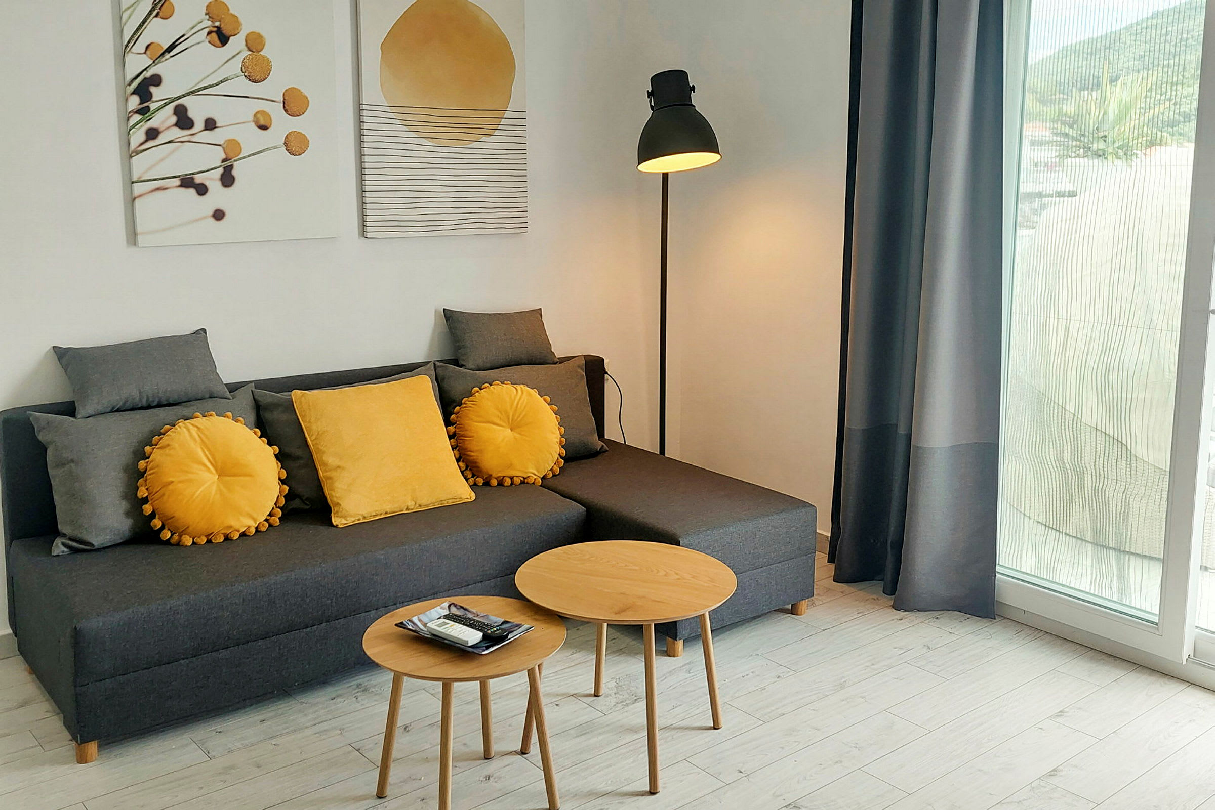 En stue med en grå sofa med gule pyntepuder, to runde træborde, en gulvlampe og vægdekorationer. Et stort vindue med gardiner kan også ses. (AI-genereret tekst)