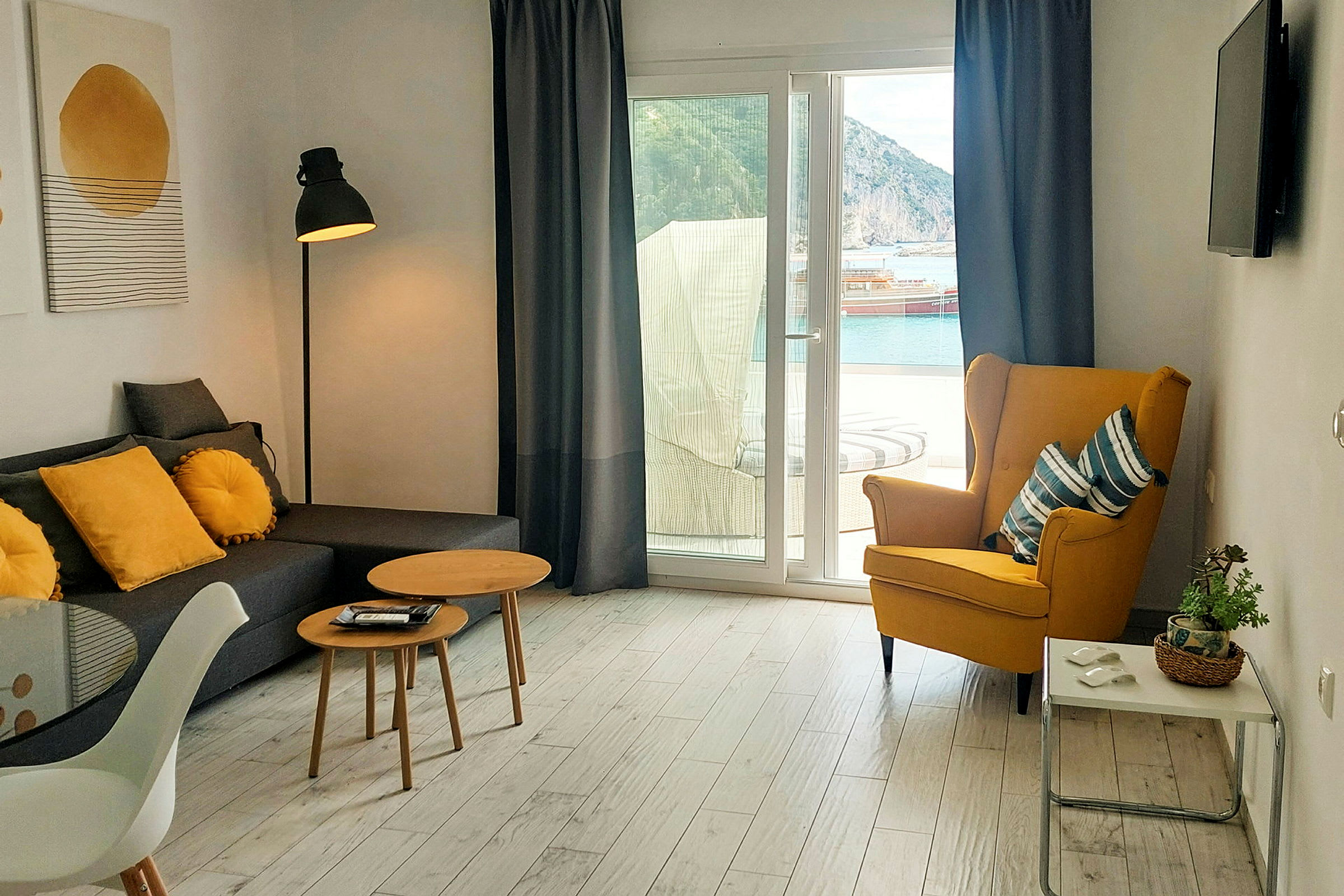 En stue med sofagruppe, gul lænestol, små borde og udsigt til en havn gennem en glasdør. (AI-genereret tekst)