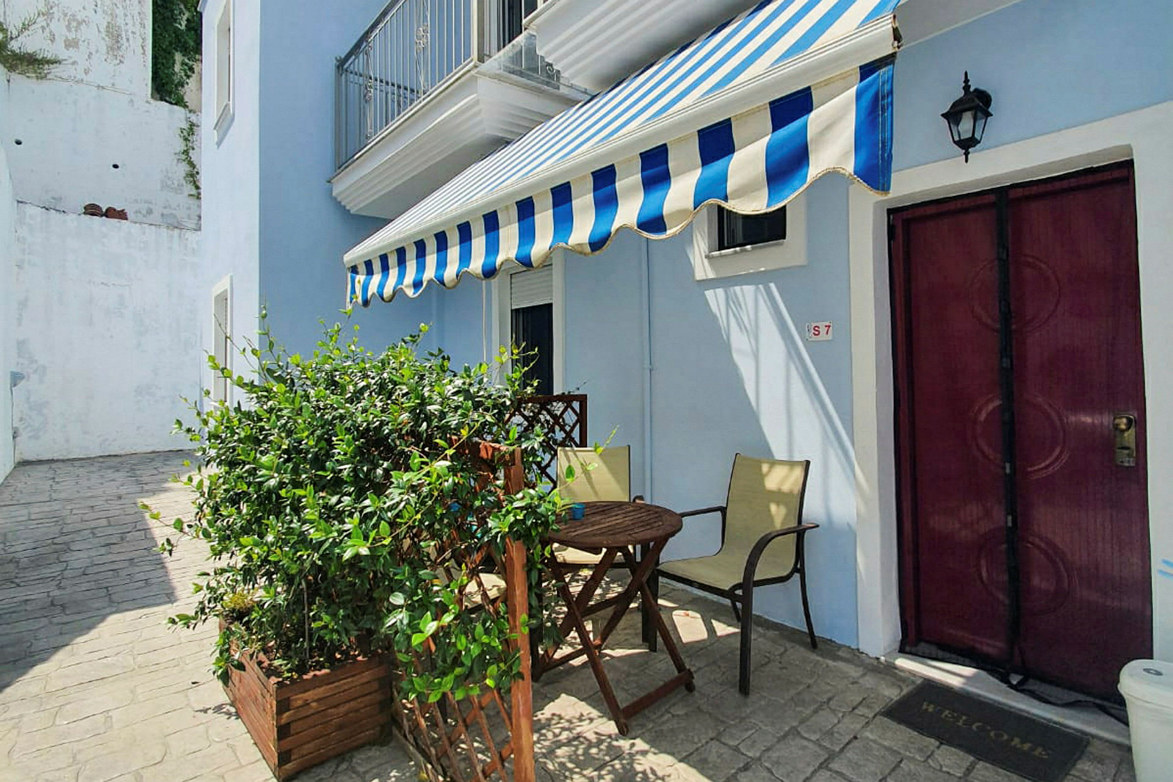 En lille udendørs terrasse med et bord, stole og en markise foran en lyseblå bygning. (AI-genereret tekst)