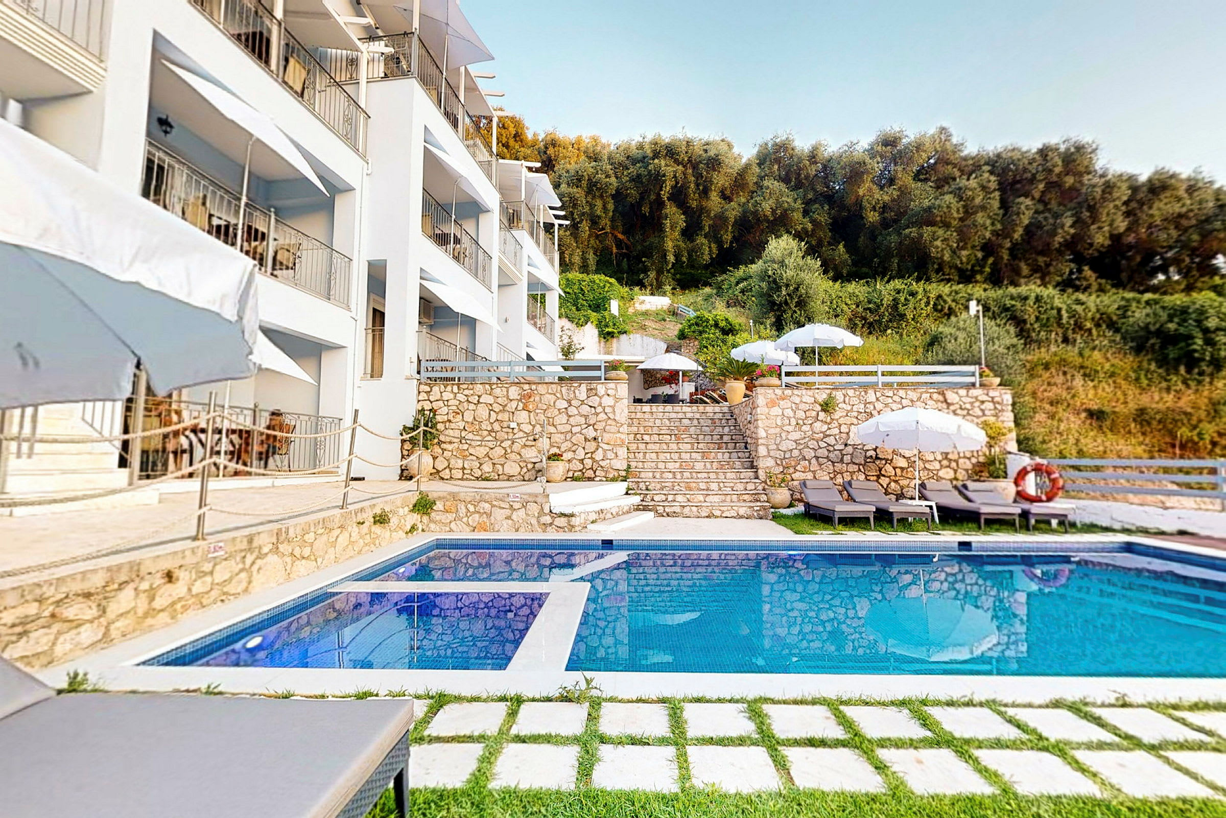 En pool foran en bygning omgivet af parasoller, liggestole og vegetation. (AI-genereret tekst)