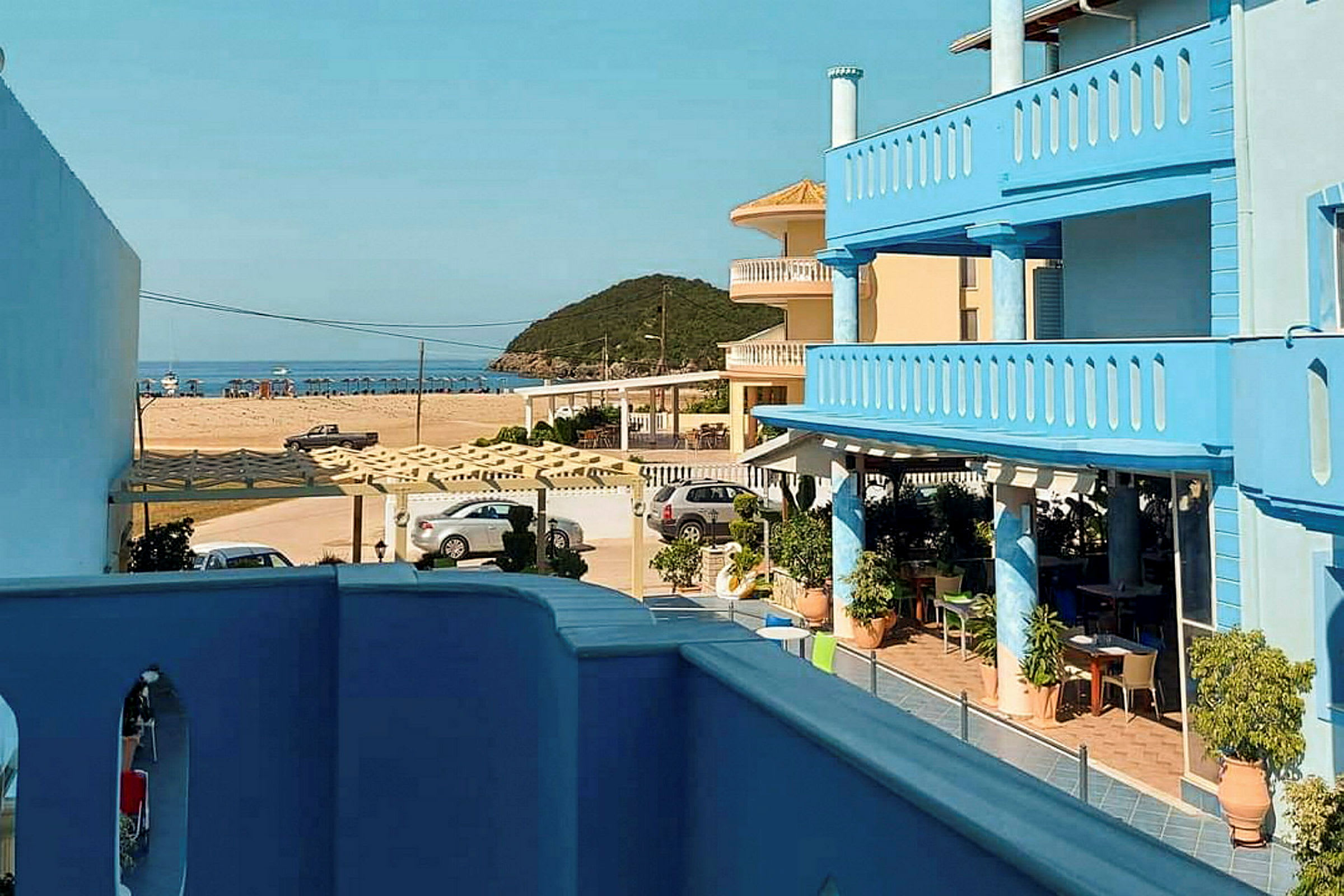 Flere bygninger med blå facader og terrasser, med udsigt mod en sandstrand og havet i baggrunden. (AI-genereret tekst)