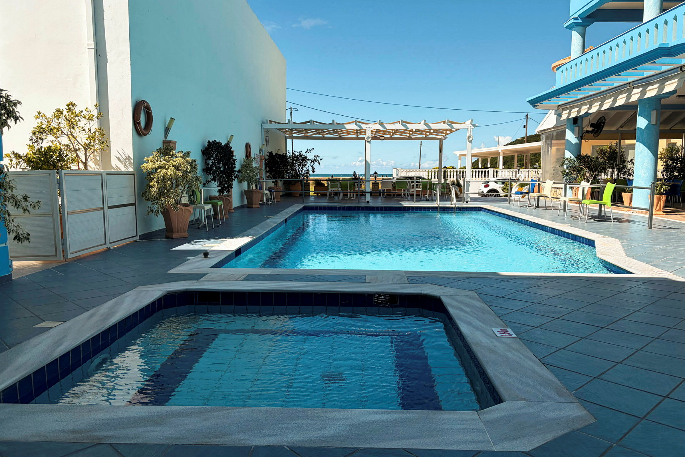 En udendørs pool med en separat jacuzzi i forgrunden, omgivet af borde, stole og planter. (AI-genereret tekst)