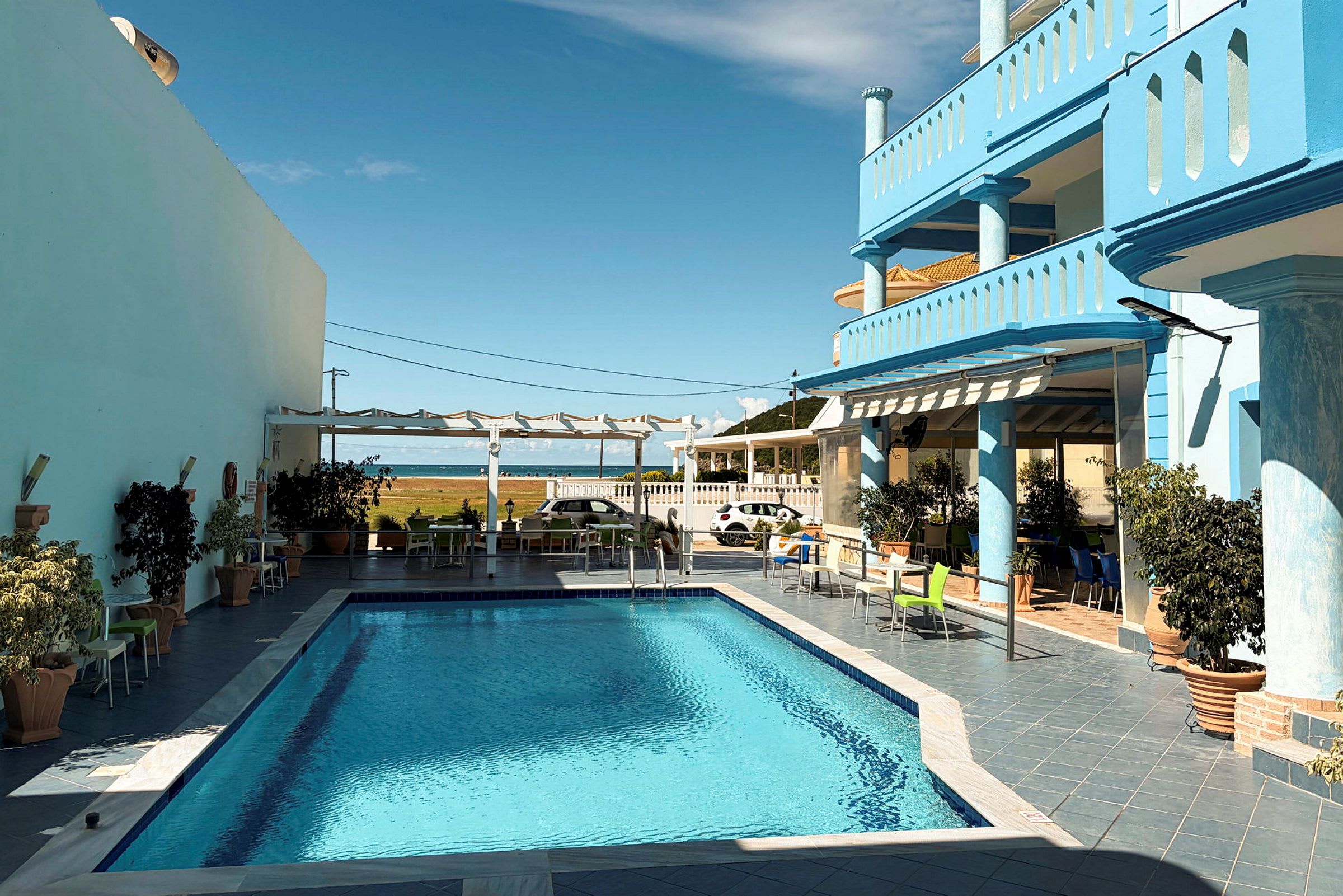 En udendørs swimmingpool omgivet af terrasse med siddepladser og potteplanter, med udsigt til havet i baggrunden. (AI-genereret tekst)