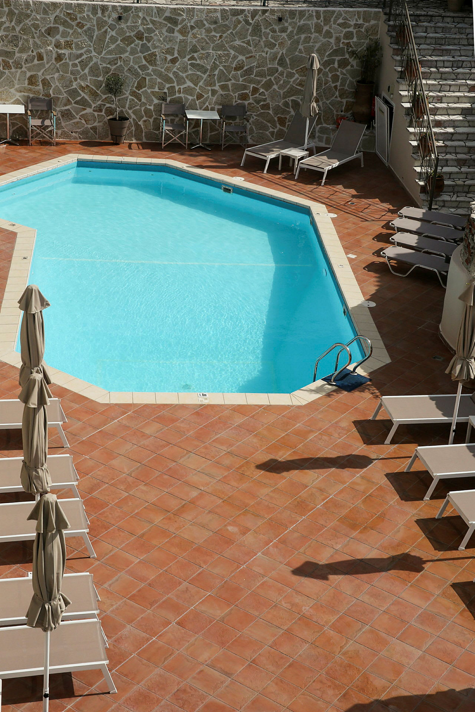 Rektangulær udendørs pool omgivet af liggestole og parasoller på terrakottafarvet gulv. (AI-genereret tekst)