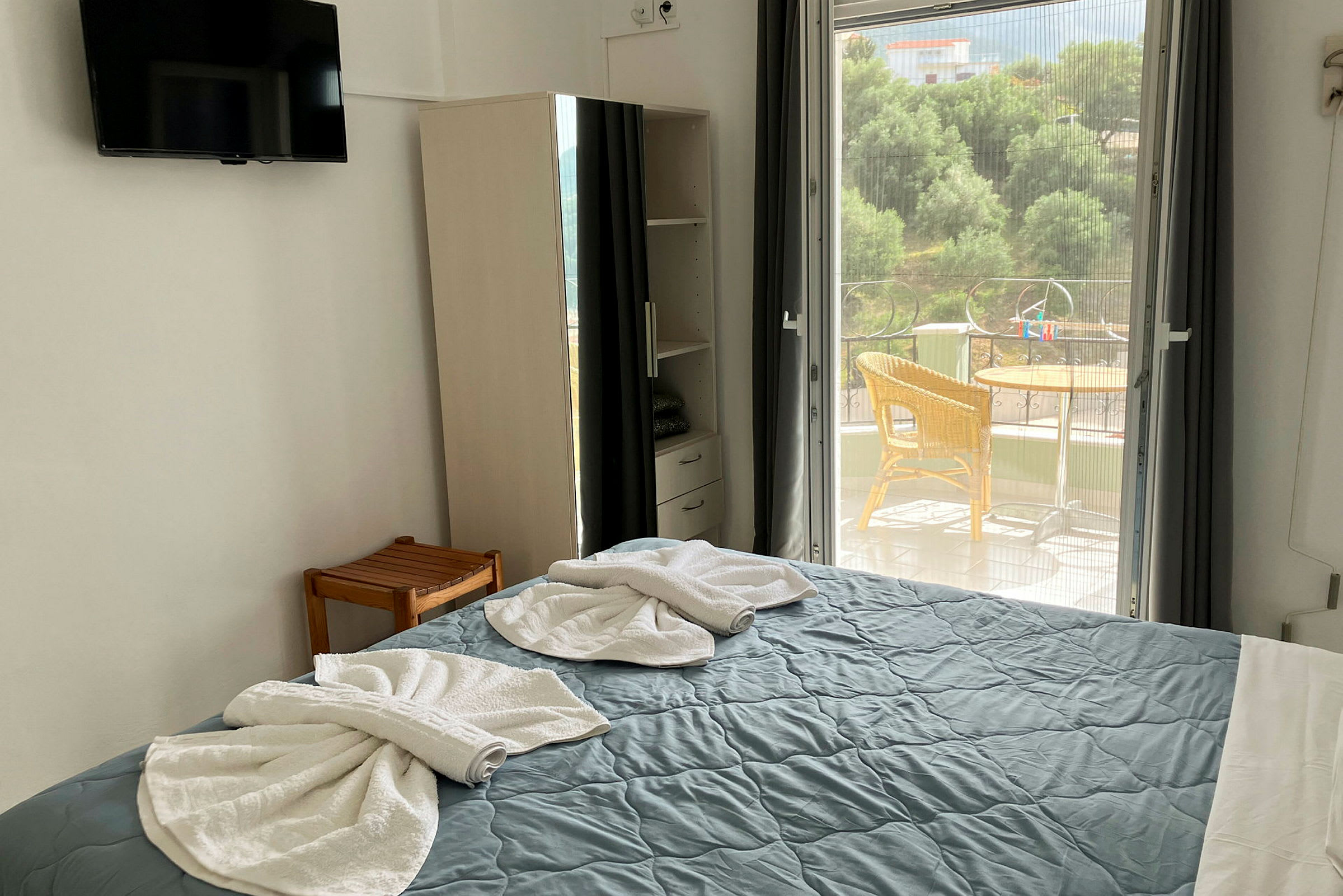 Et hotelværelse viser en opredt seng med foldede håndklæder, et skab og en udgang til en balkon. (AI-genereret tekst)