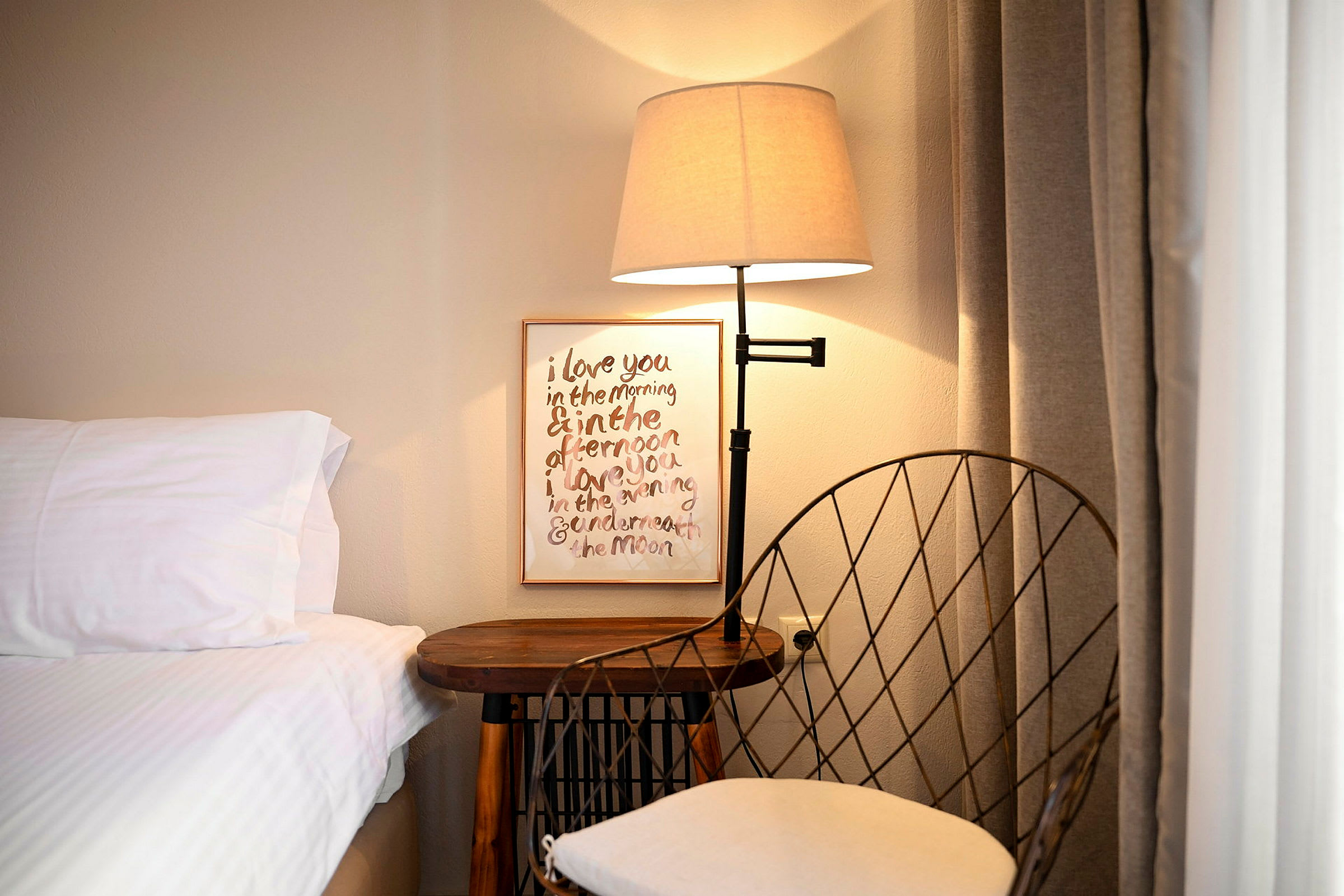 Et natbord med en lampe og en stol ved siden af en seng. En tavle med tekst hænger på væggen. (AI-genereret tekst)