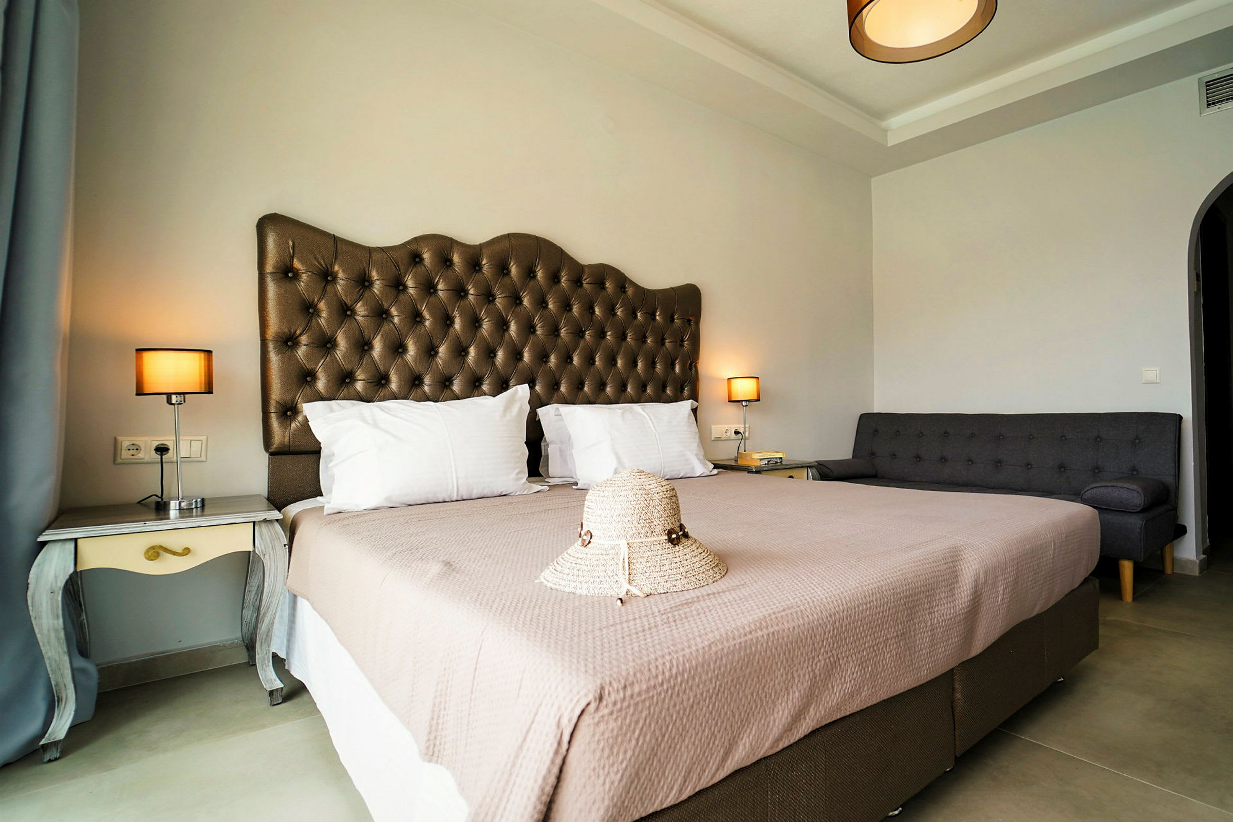 Et hotelværelse med en dobbeltseng, en sofa og natborde med lamper, samt en hat placeret på sengen. (AI-genereret tekst)