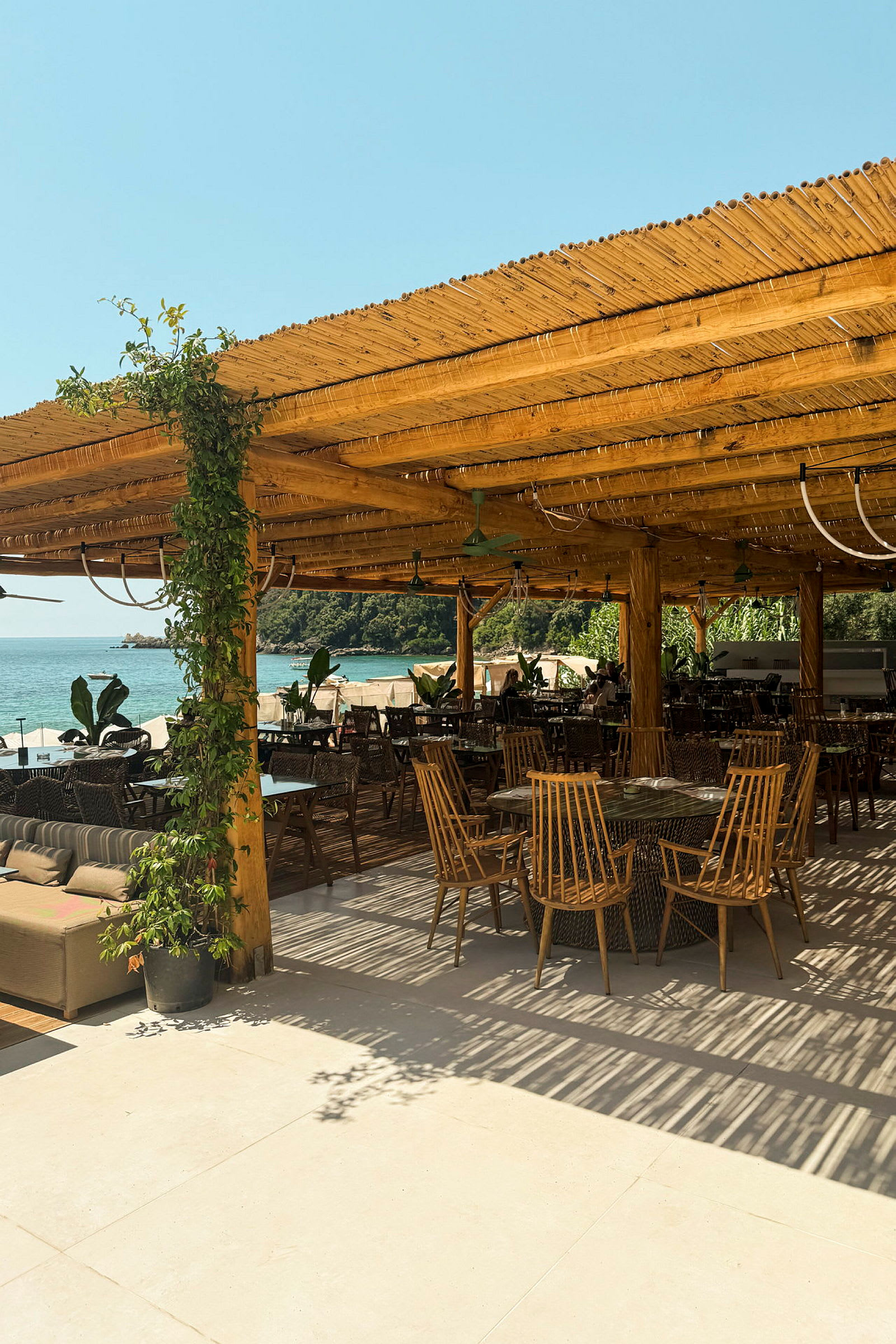 Udendørs serveringsområde med træpergola, borde og stole samt udsigt til havet. (AI-genereret tekst)