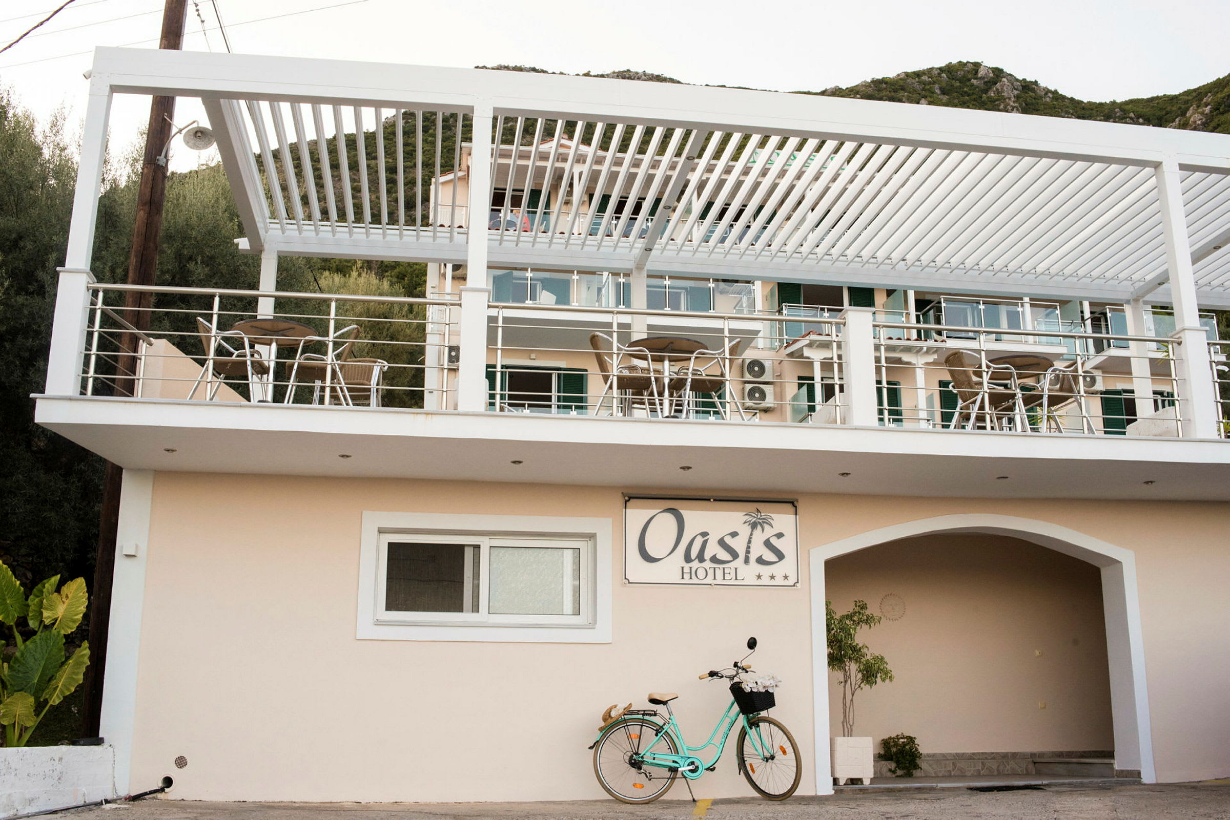 Billede af Oasis Hotel med altaner og udemøbler samt en cykel parkeret nedenfor. (AI-genereret tekst)