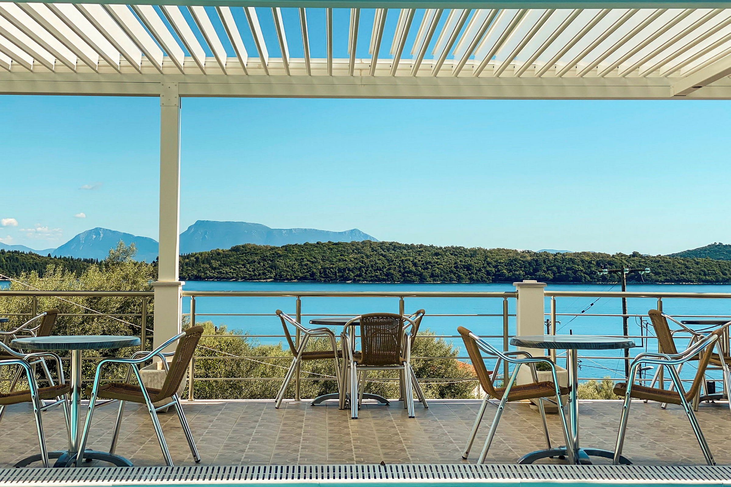 Udendørs møbler med borde og stole på en terrasse med udsigt over havet og skovklædte bakker. (AI-genereret tekst)
