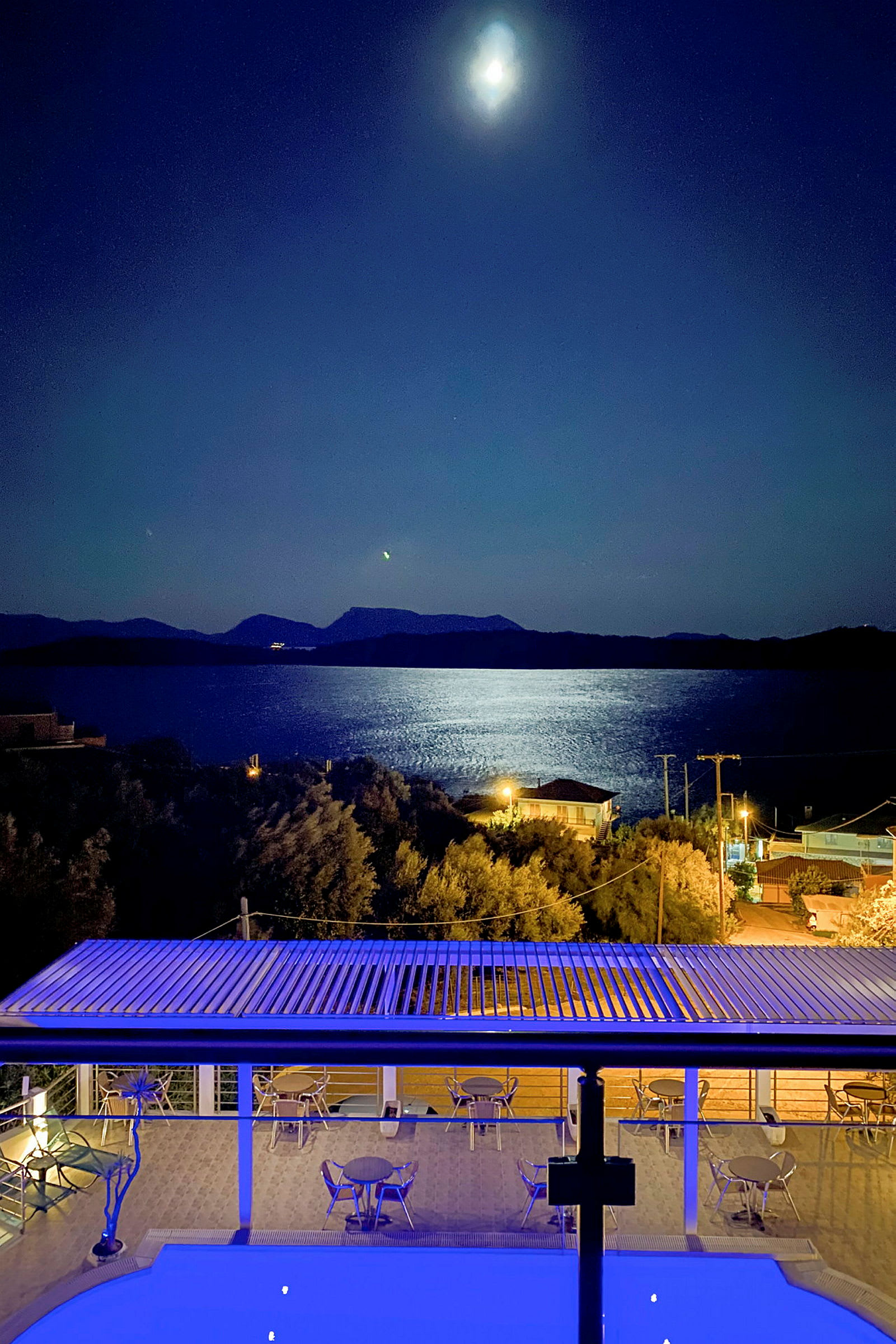 Natudsigt over en møbleret terrasse, pool og hav med måneskin. (AI-genereret tekst)