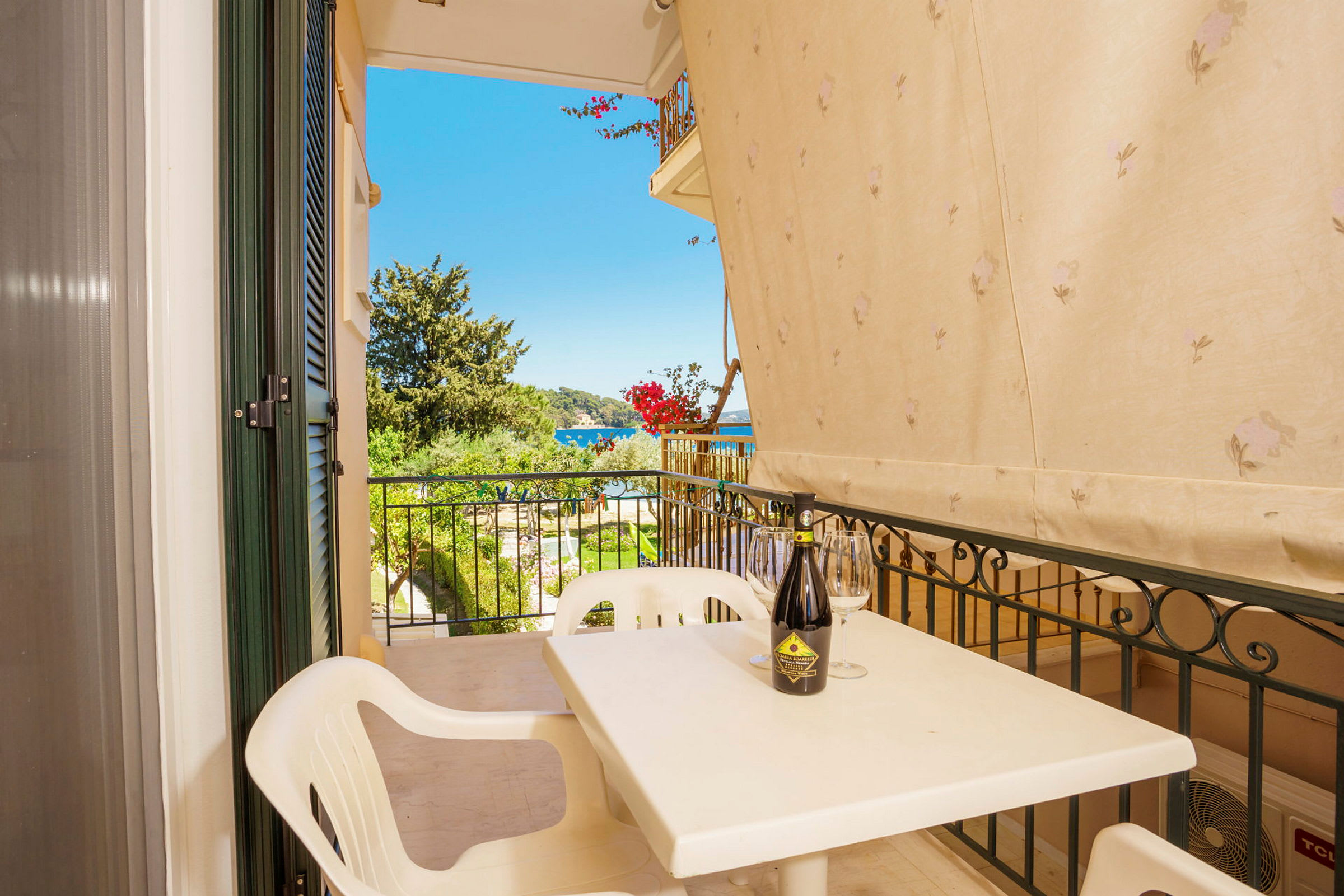 Udsigt fra en balkon med en plastikstol, et bord med en flaske og glas, samt omgivelser med træer og hav i det fjerne. (AI-genereret tekst)