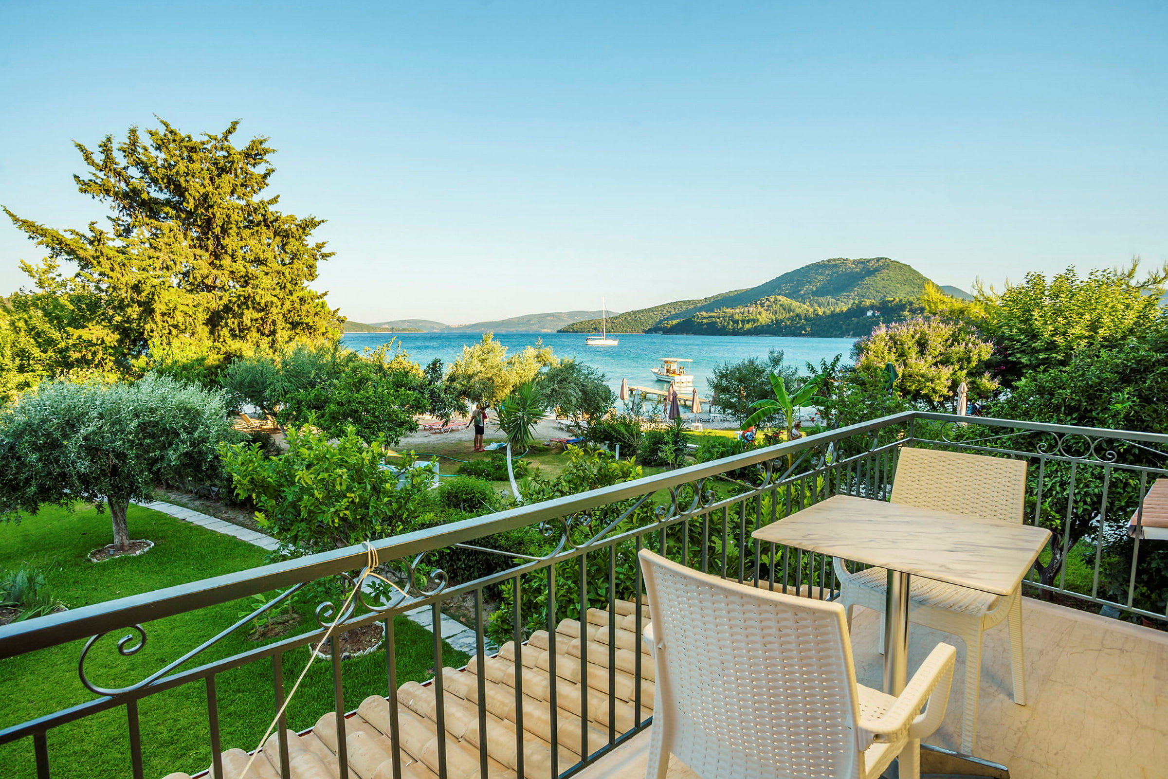 En balkon med bord og stole, udsigt over en grøn have, havet og bjerge i baggrunden. (AI-genereret tekst)