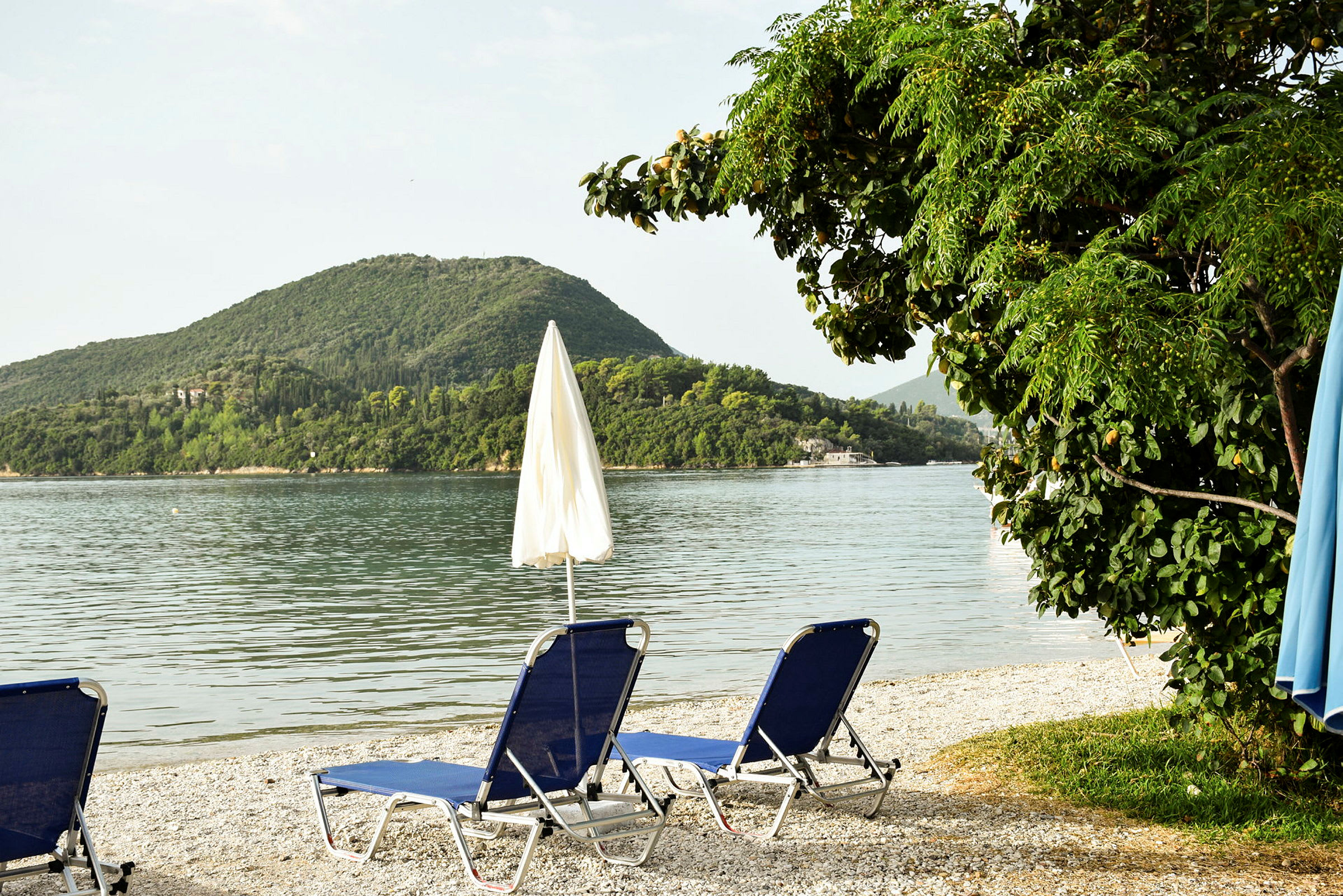 To blå solsenge med en parasol på en stenstrand ved en rolig sø, omgivet af grønne bakker. (AI-genereret tekst)