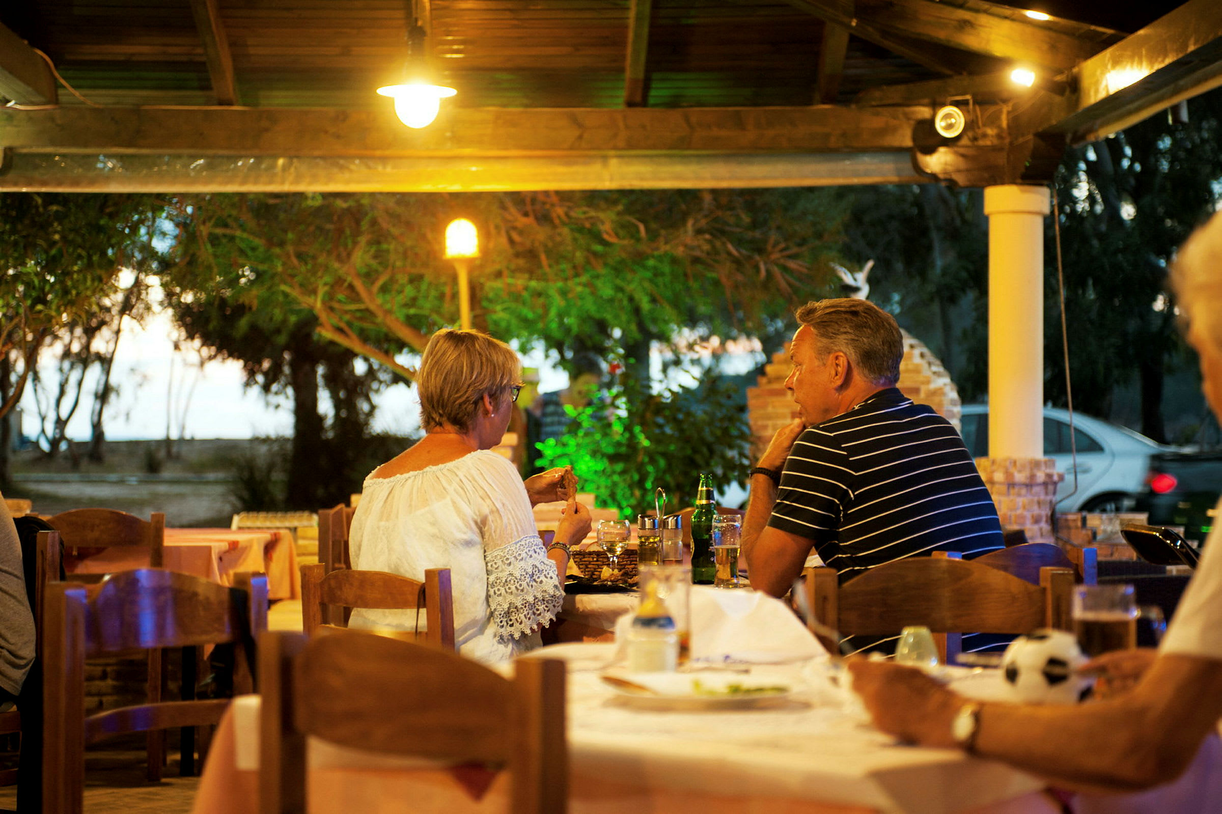 Personer sidder ved et bord på en udendørs restaurant omgivet af grønne omgivelser. (AI-genereret tekst)