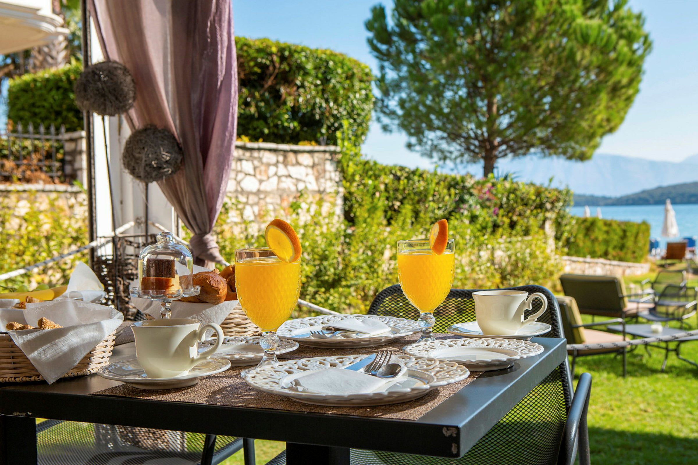 Et bord med morgenmad servering, herunder glas med juice pyntet med appelsinskiver, kopper og tallerkener, ved en have tæt på havet. (AI-genereret tekst)