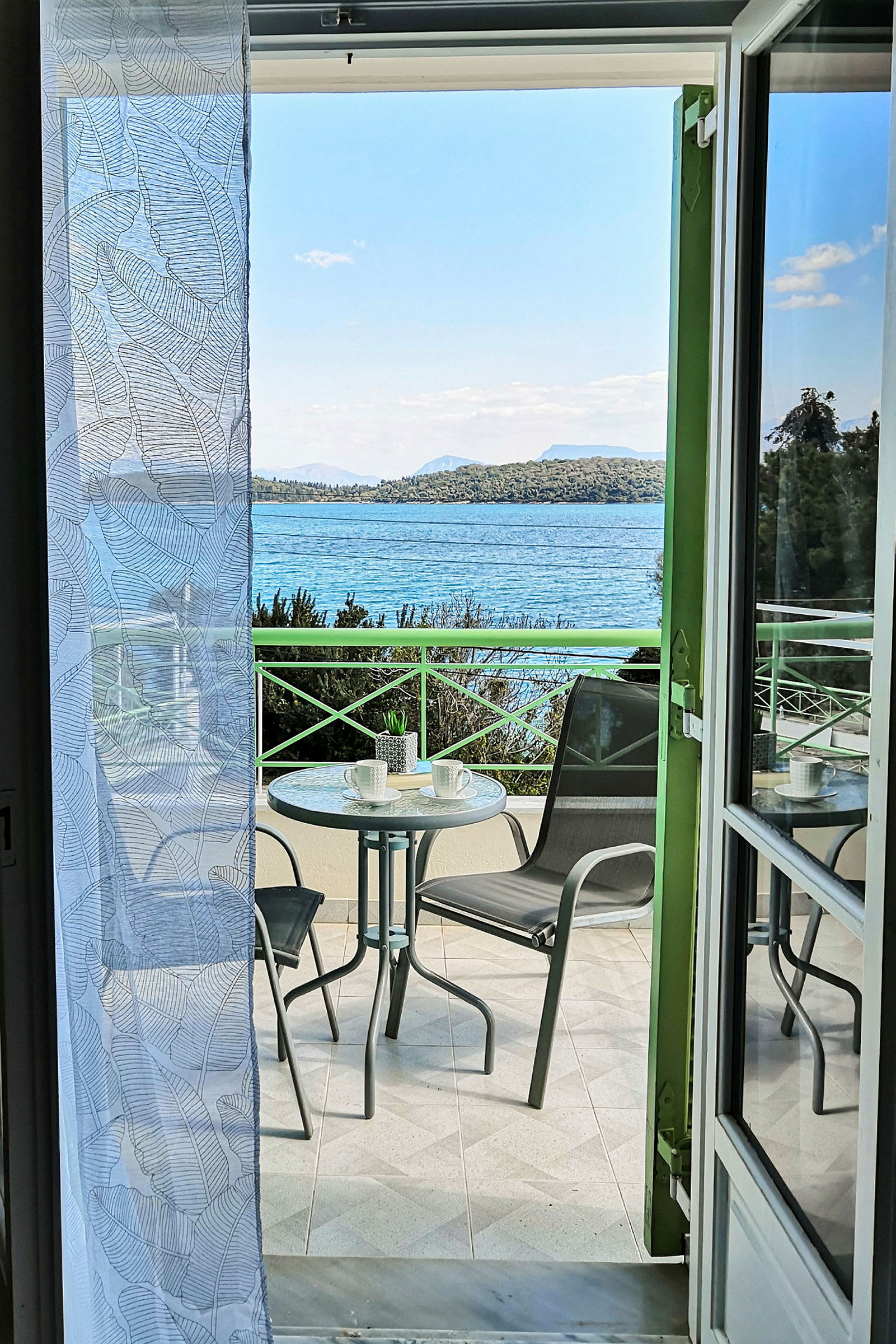 Udsigt fra et værelse mod en balkon med et bord og stole, med havet og landskabet i baggrunden. (AI-genereret tekst)