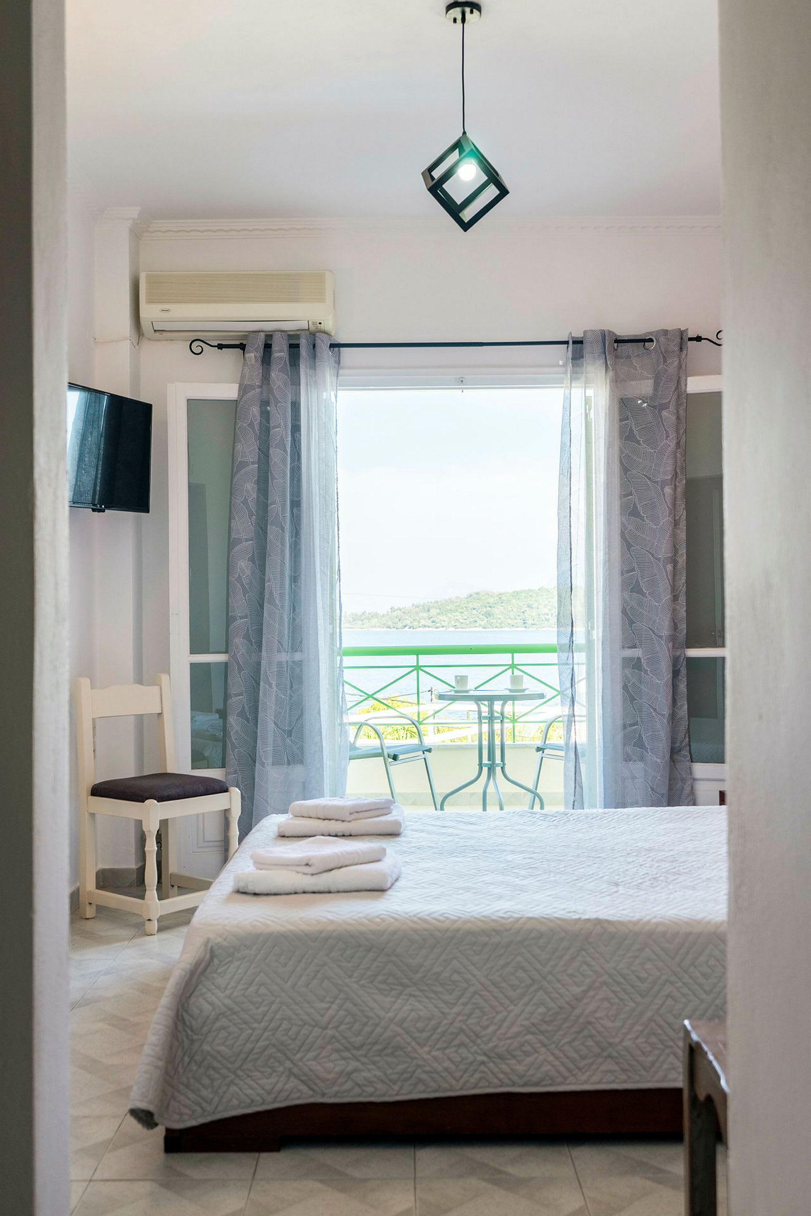 Et soveværelse med en seng med hvide sengelinned, en stol og et glasvindue, der fører ud til en balkon med bord og stole, udsigt til grønne områder og vand. (AI-genereret tekst)