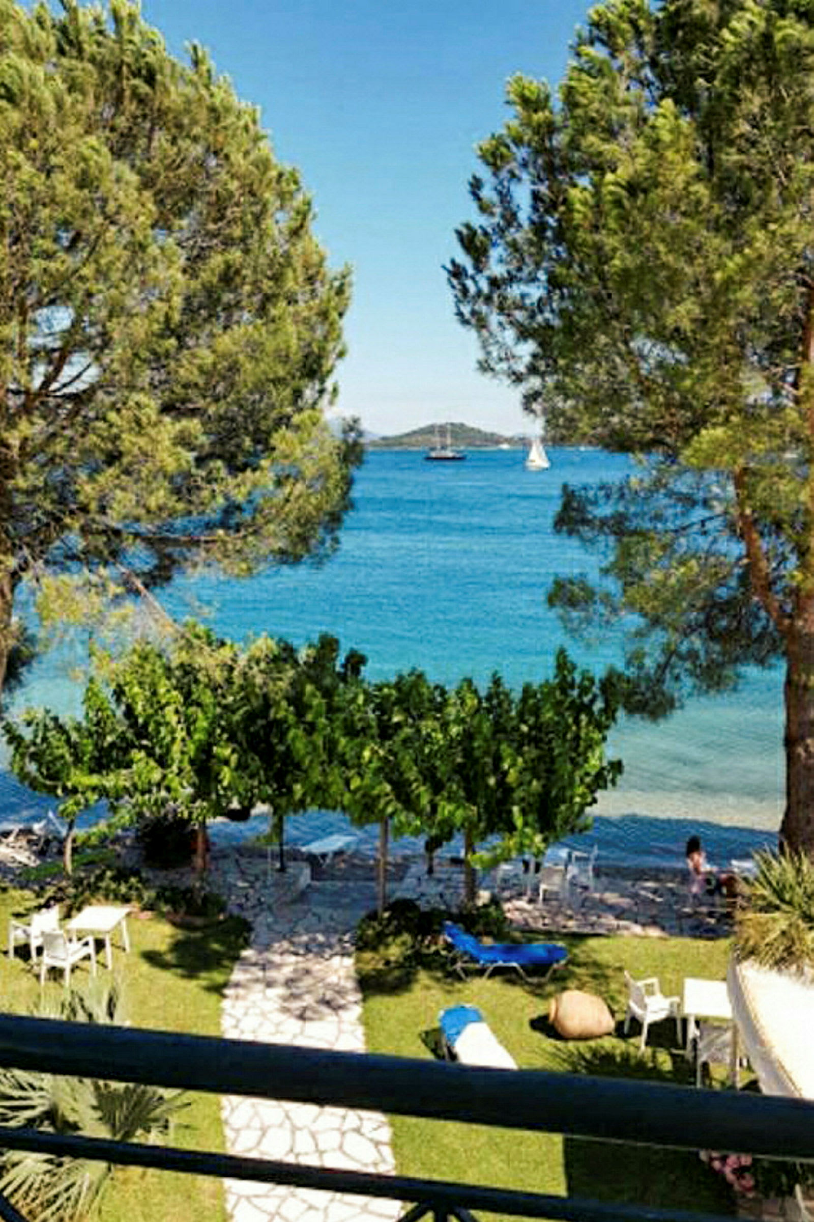 Udsigt fra en terrasse med træer, græsplæne, stole og borde, og havet i baggrunden. (AI-genereret tekst)