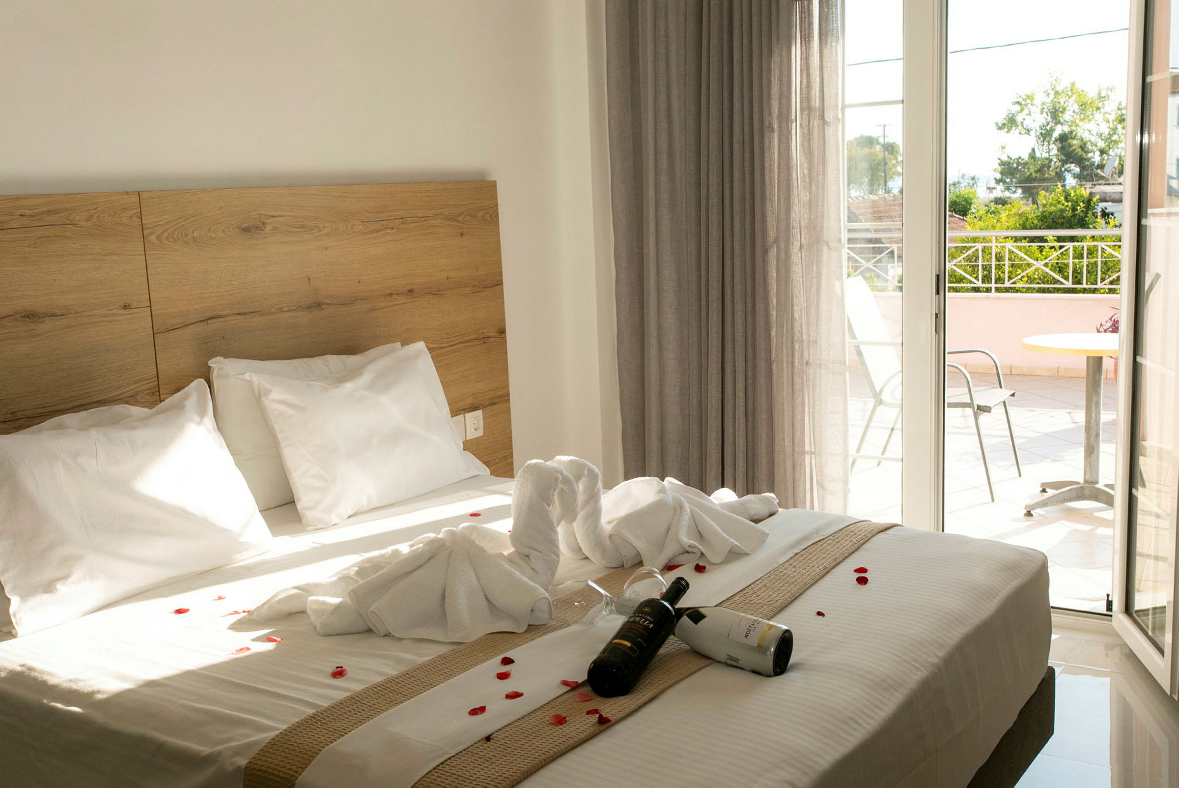 Hotelværelse med en dobbeltseng dekoreret med håndklæder og blomsterblade. En flaske vin og glas ligger på sengen. En balkon er synlig i baggrunden. (AI-genereret tekst)