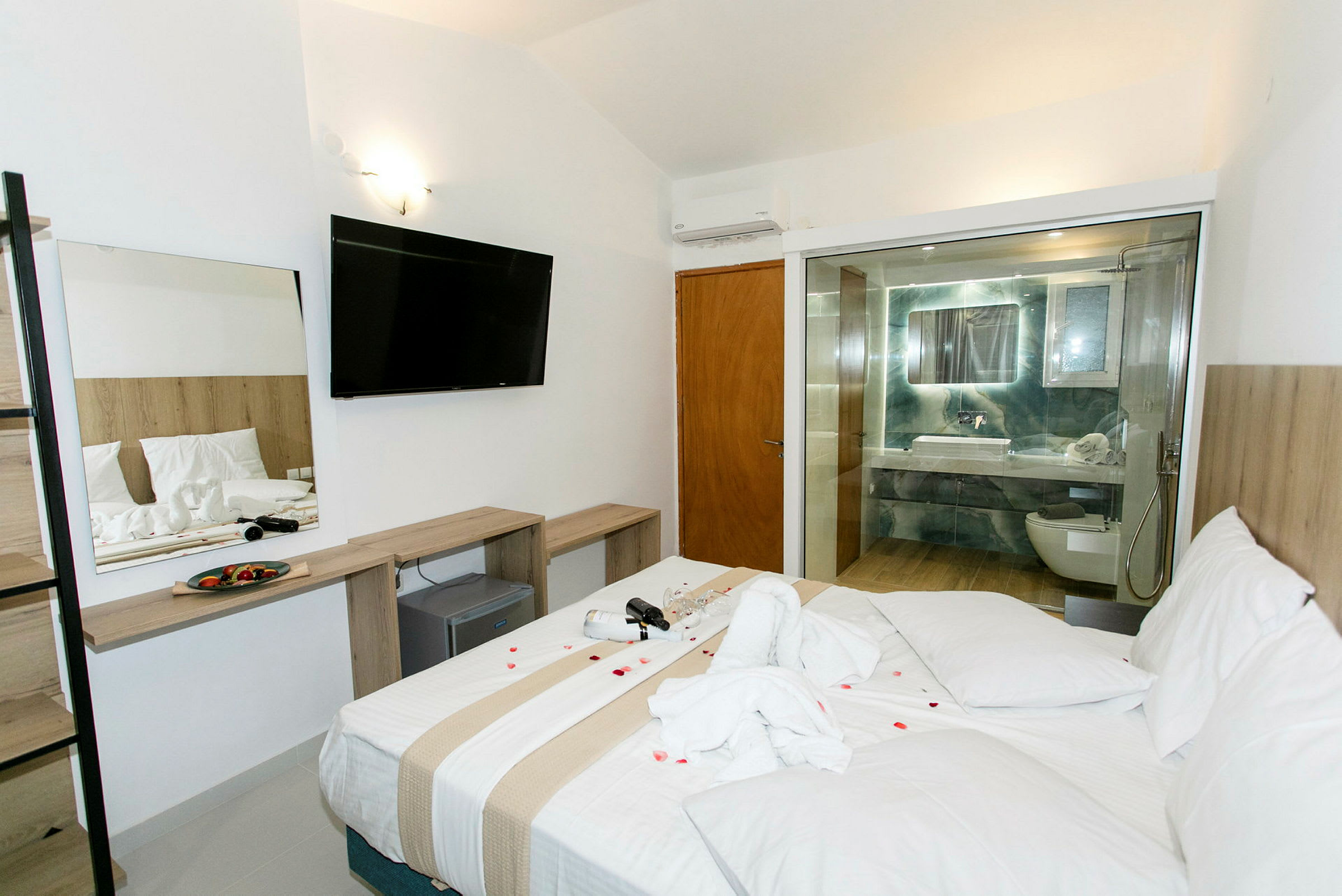 Hotelværelse med en stor seng, et åbent badeværelse med glasvæg og en møblering inklusive et tv. (AI-genereret tekst)