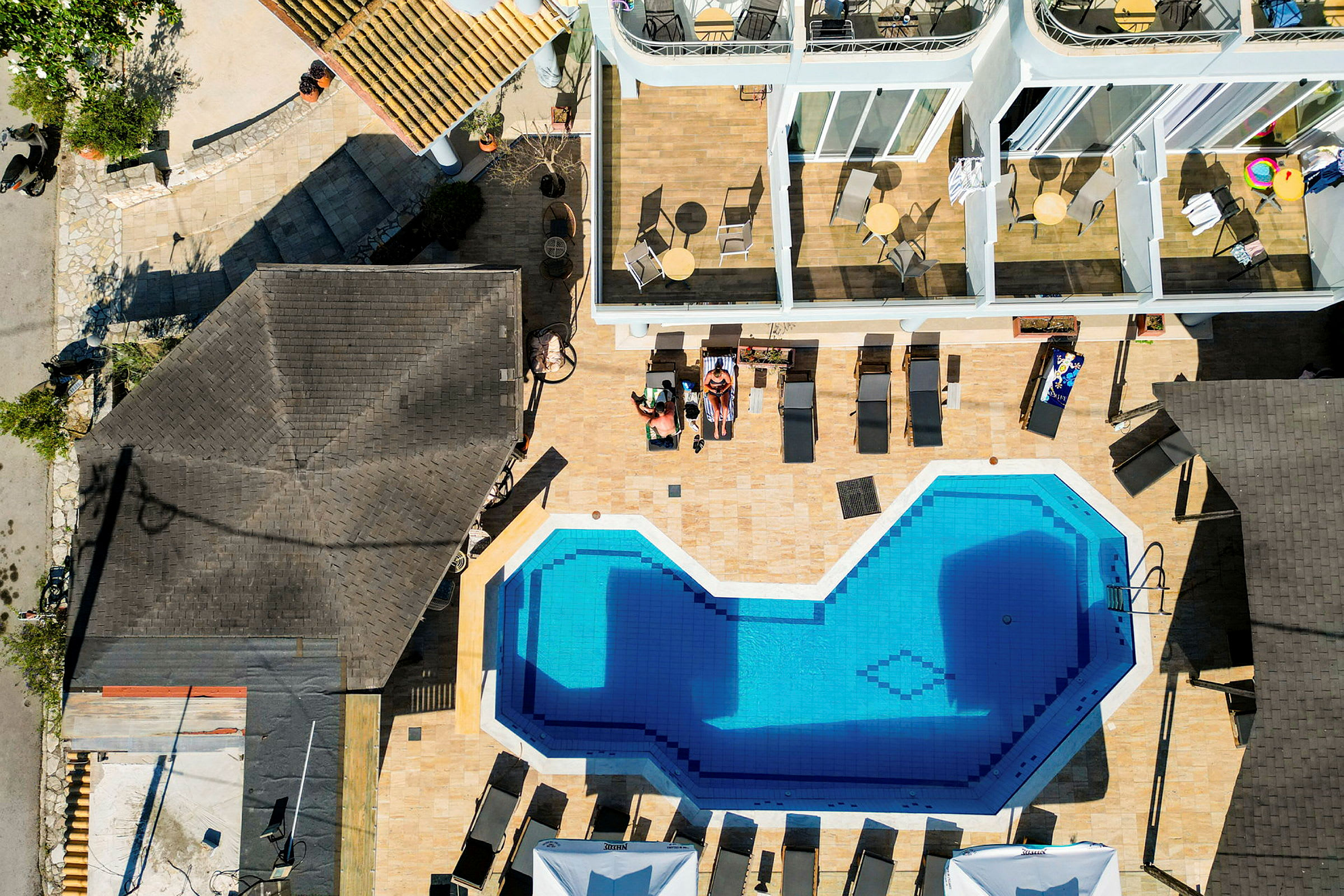 Dronebillede af en swimmingpool omgivet af liggestole og bygninger med terrasser. (AI-genereret tekst)