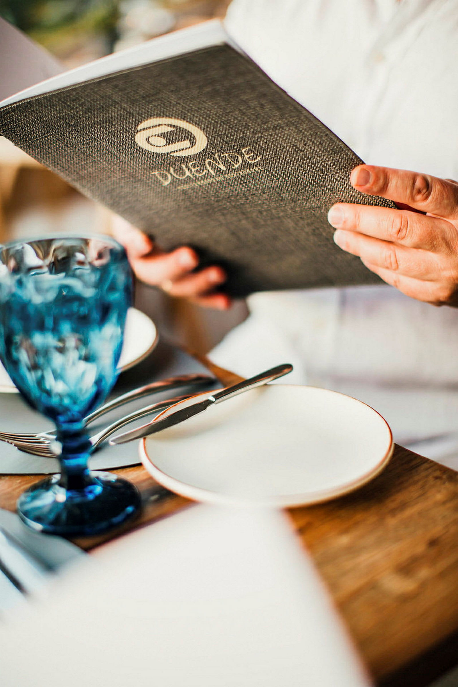 En person holder en menu ved et dækket bord med et blåt drikkeglas. (AI-genereret tekst)