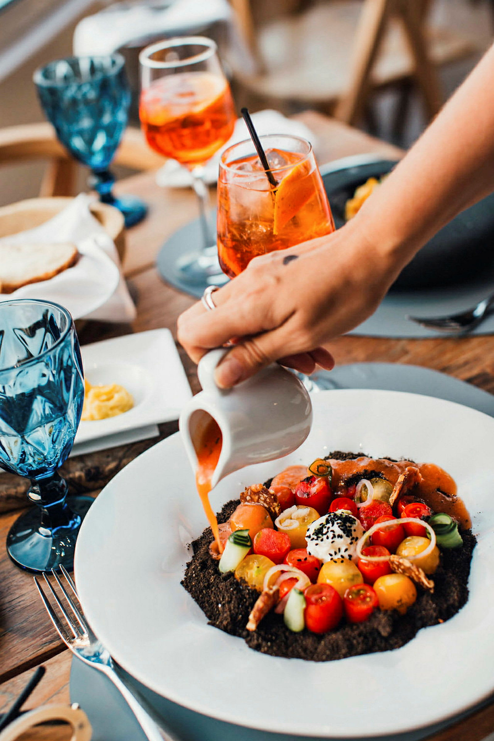 En person hælder sovs fra en lille kande over en gourmetret på en tallerken, omgivet af glas og andet tilbehør på et bord. (AI-genereret tekst)