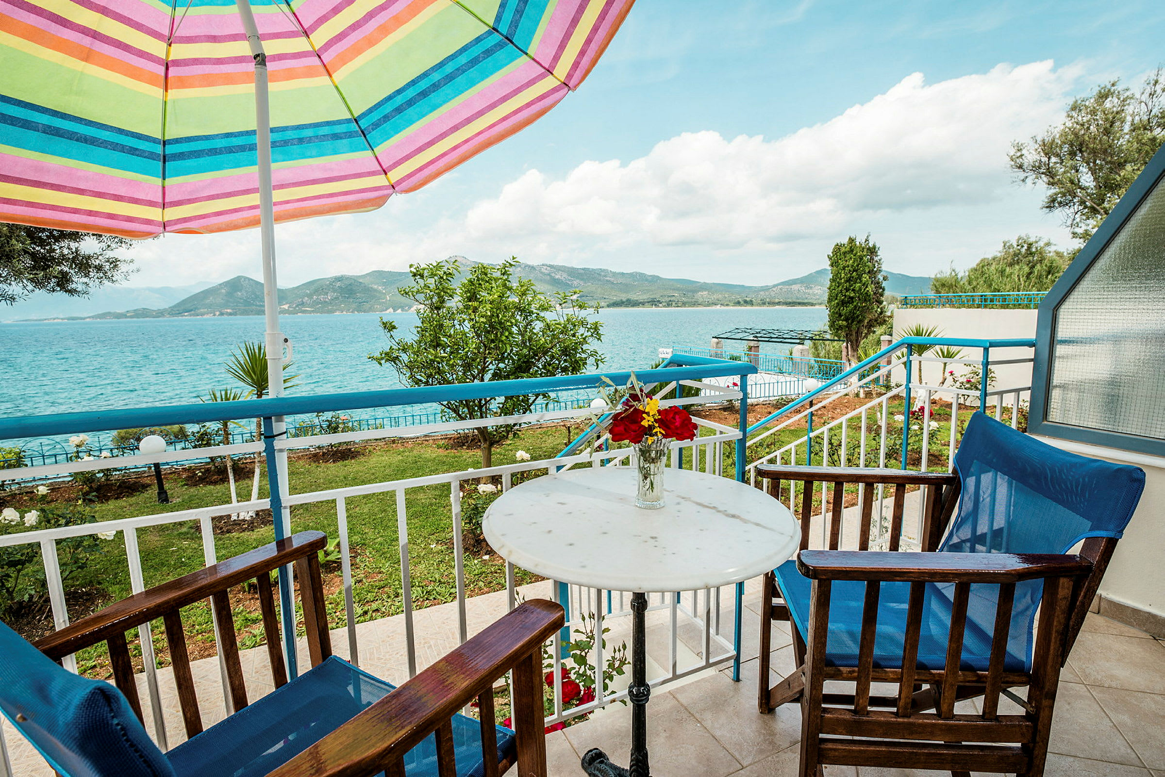 Udendørs område med bord, stole og en farverig parasol, med udsigt over havet og landskabet. (AI-genereret tekst)