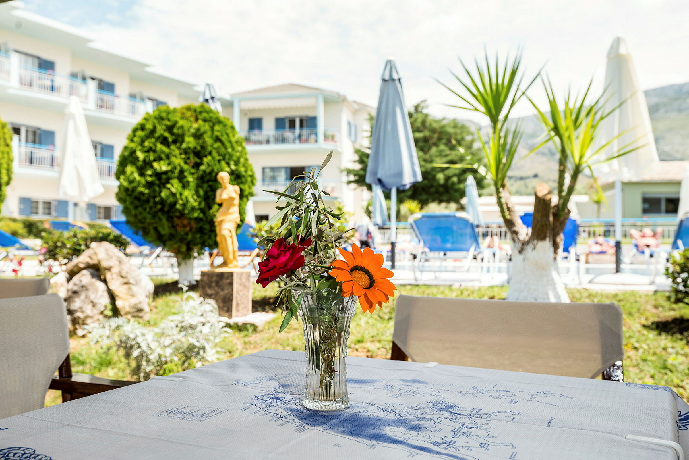 Billede af et bord med en vase med blomster, i baggrunden ses en bygning, parasoller og et poolområde. (AI-genereret tekst)