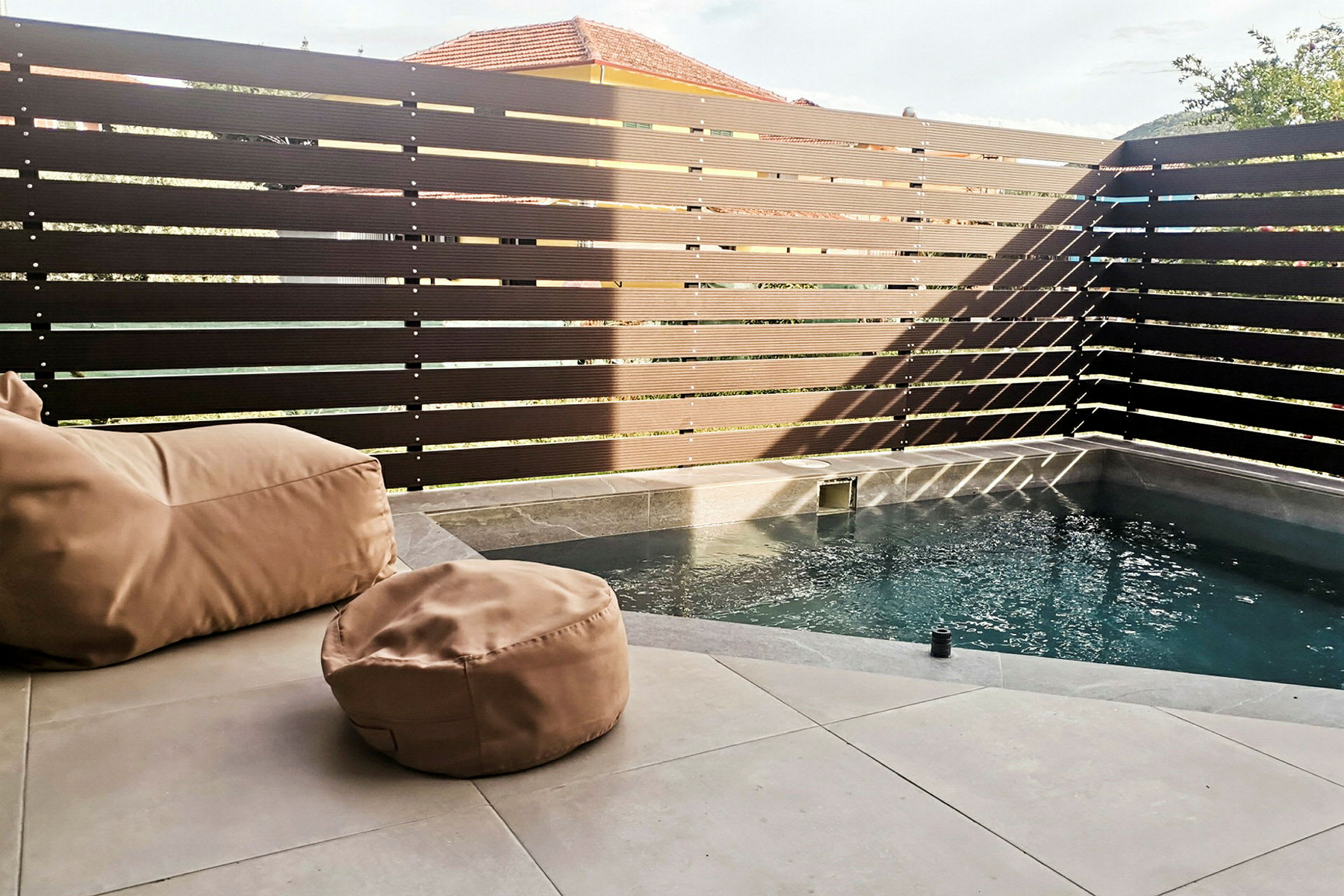 Lille pool med beige sækkestole på en stenoverflade, omgivet af et højt, mørkt hegn. (AI-genereret tekst)