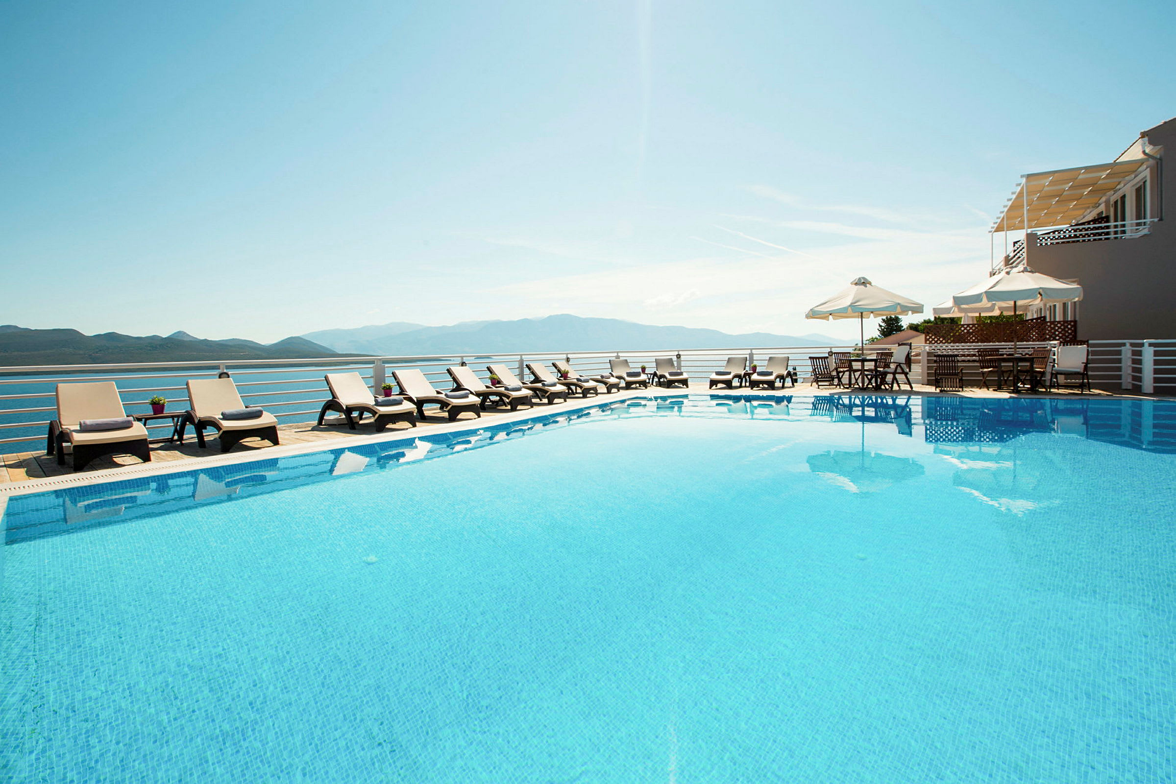 En udendørs swimmingpool omgivet af liggestole og parasoller. Udsigt over bjerge og himmel i baggrunden. (AI-genereret tekst)