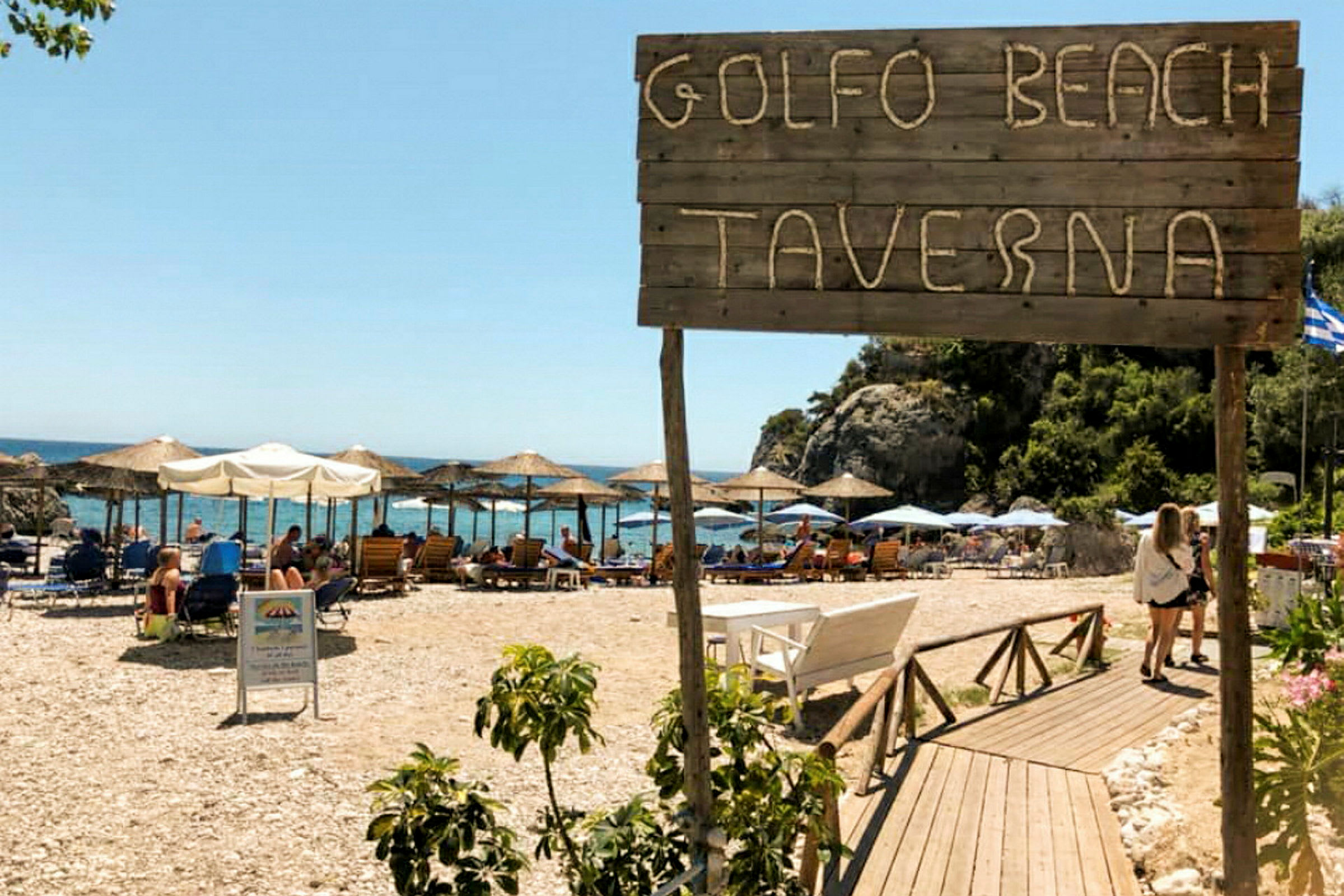 Skilt med teksten 'Golfo Beach Taverna' foran en strand med parasoller og liggestole. (AI-genereret tekst)